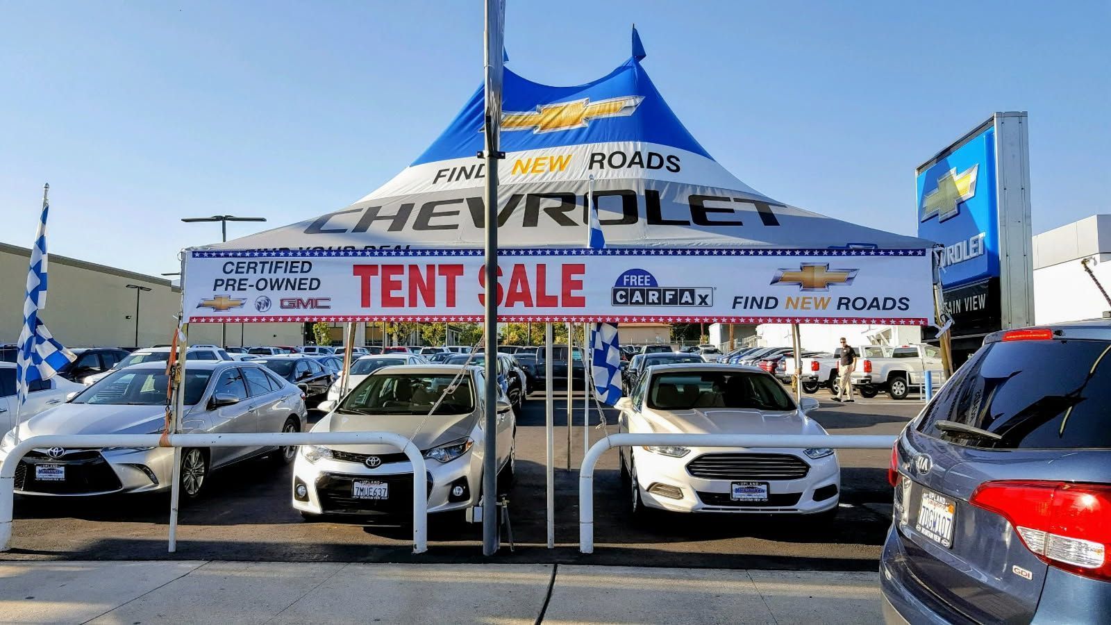 Car dealership tent sale with several parked cars and red “TENT SALE” banners under a blue canopy.