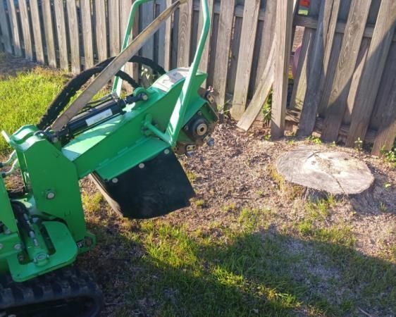 A green stump grinder is sitting in front of a wooden fence.