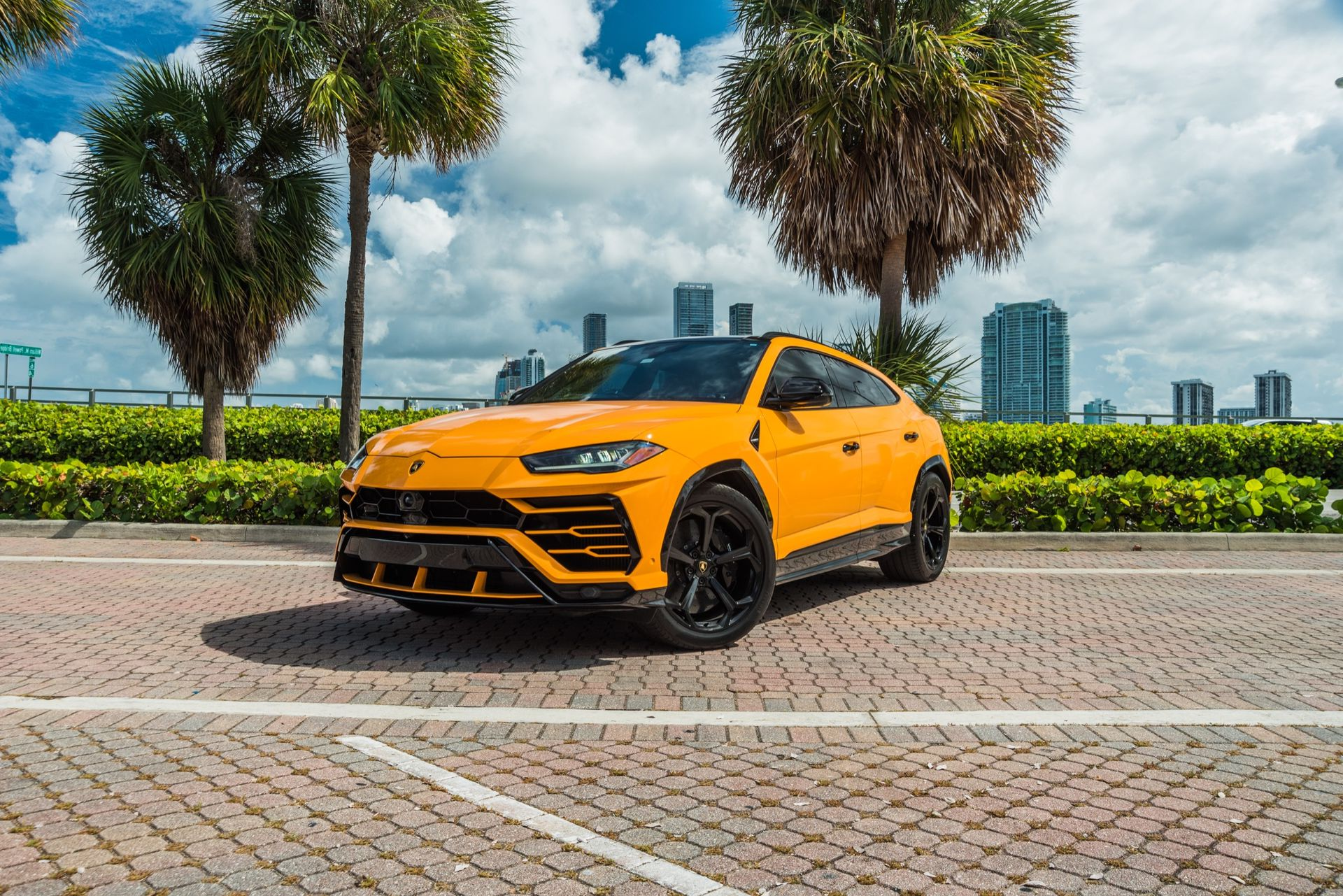 Yellow Lamborghini SUV parked on brick pavement in Miami, with palm trees and city skyline.