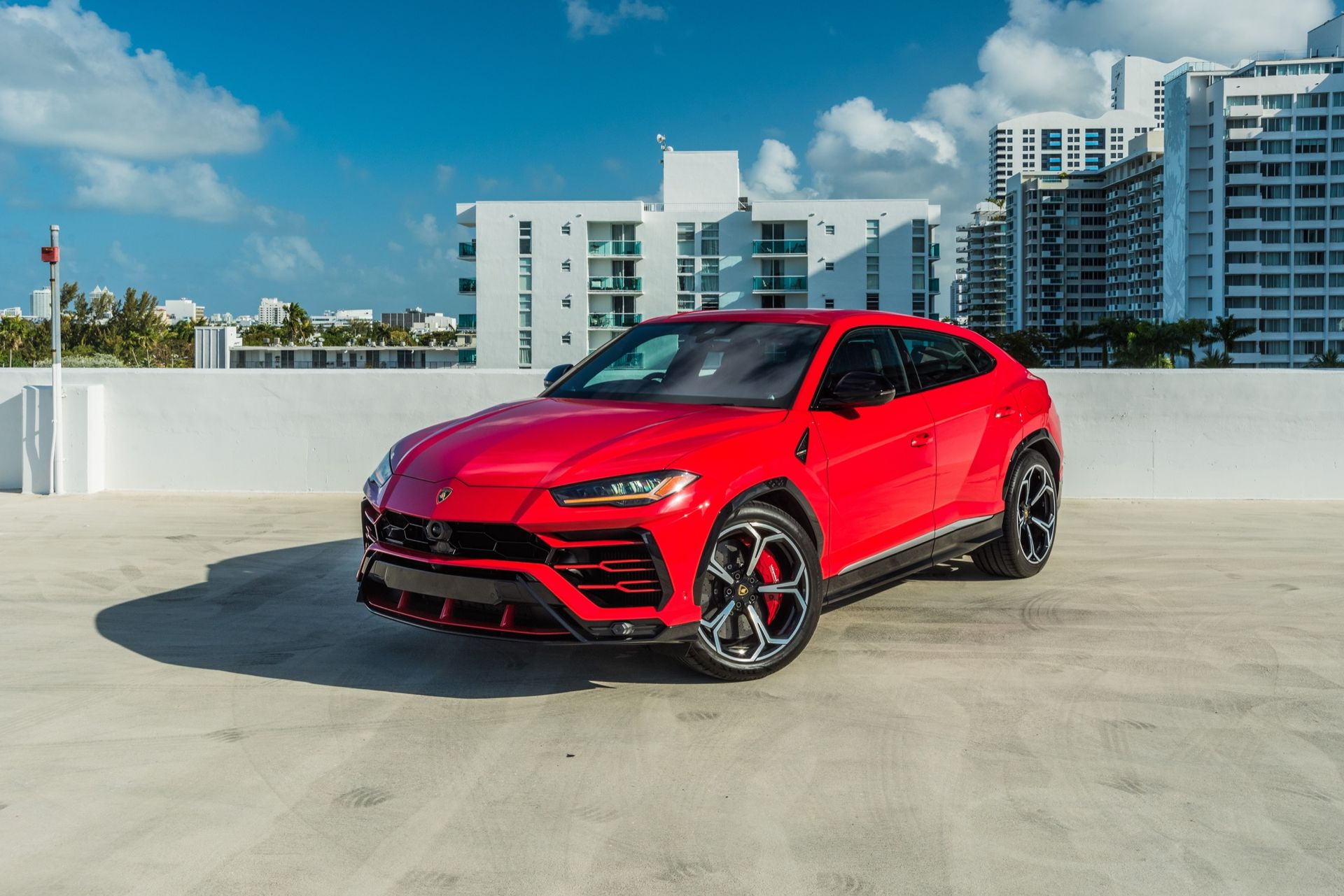 Red Lamborghini SUV parked on a concrete rooftop with a city backdrop on a sunny day.