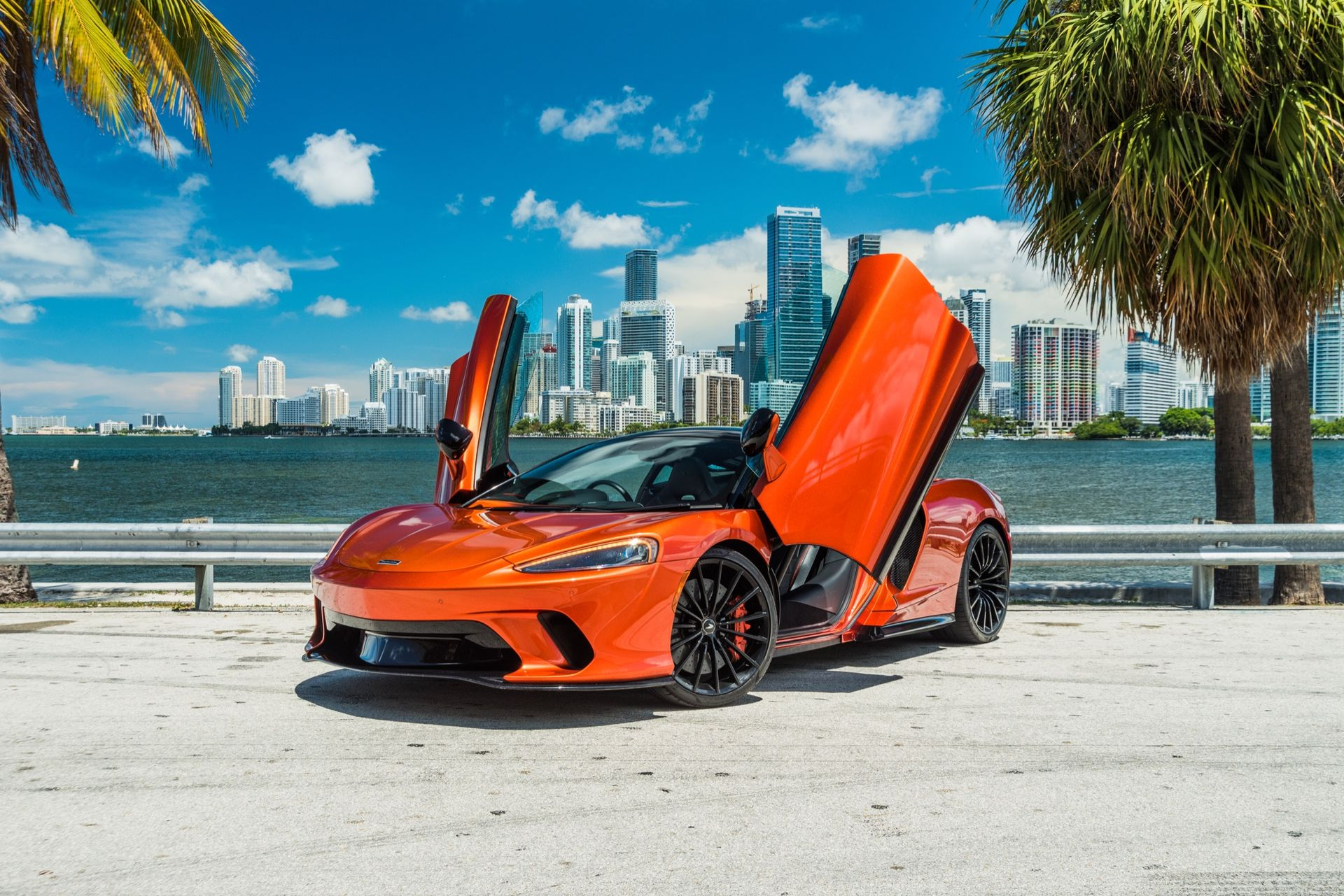 Orange sports car with open doors parked in front of a waterfront, city skyline backdrop.