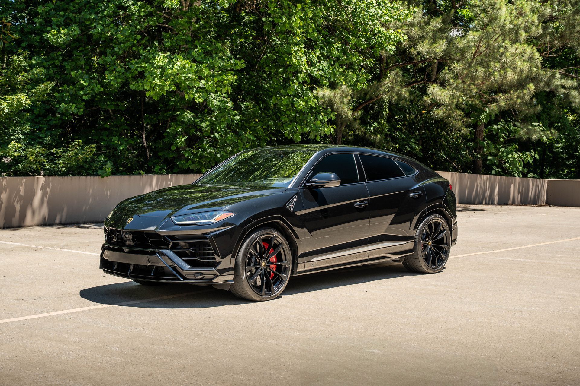 Black Lamborghini Urus SUV parked on asphalt, green trees in background.