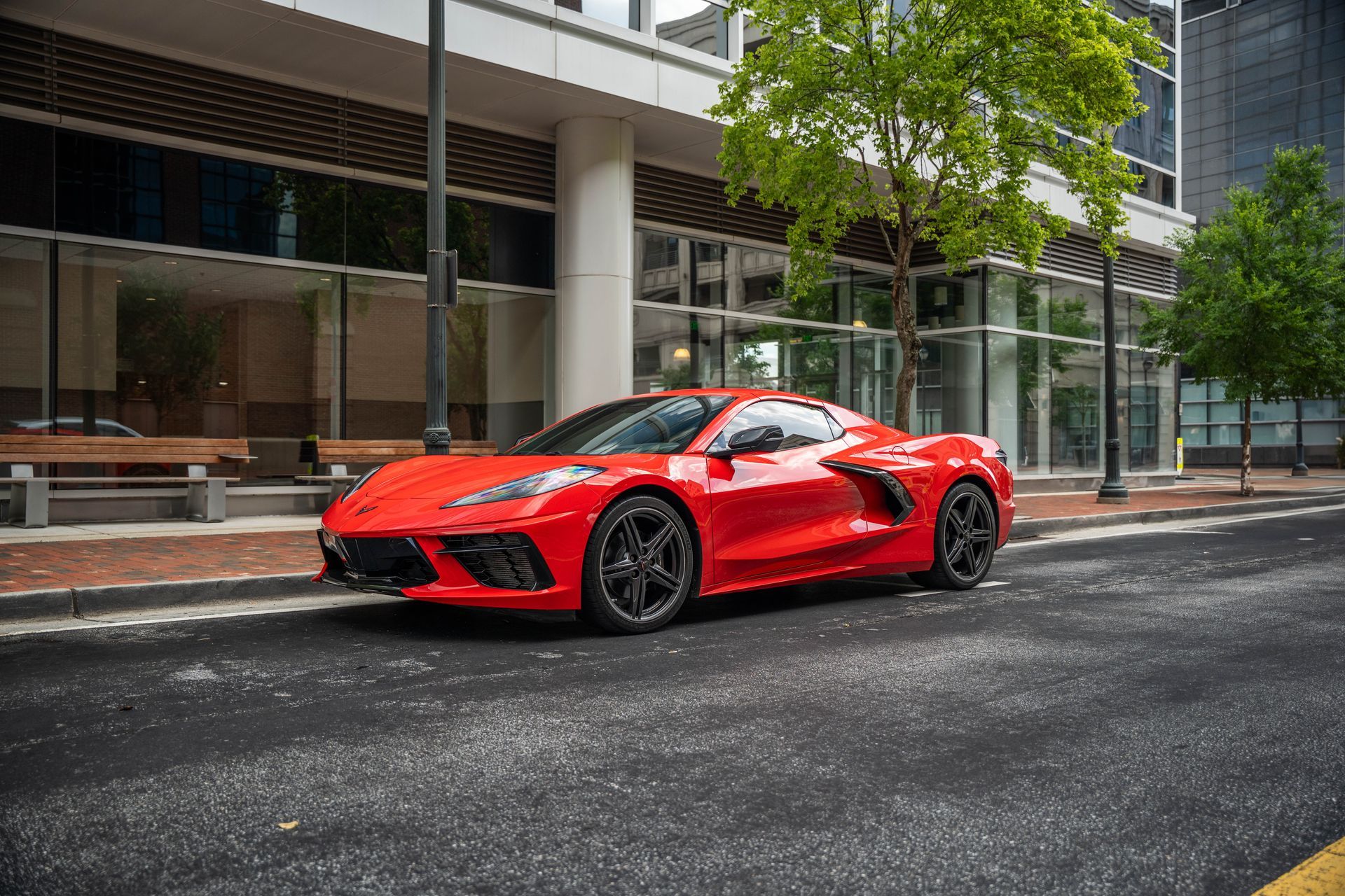 Red sports car parked on a city street in front of a modern building and trees.