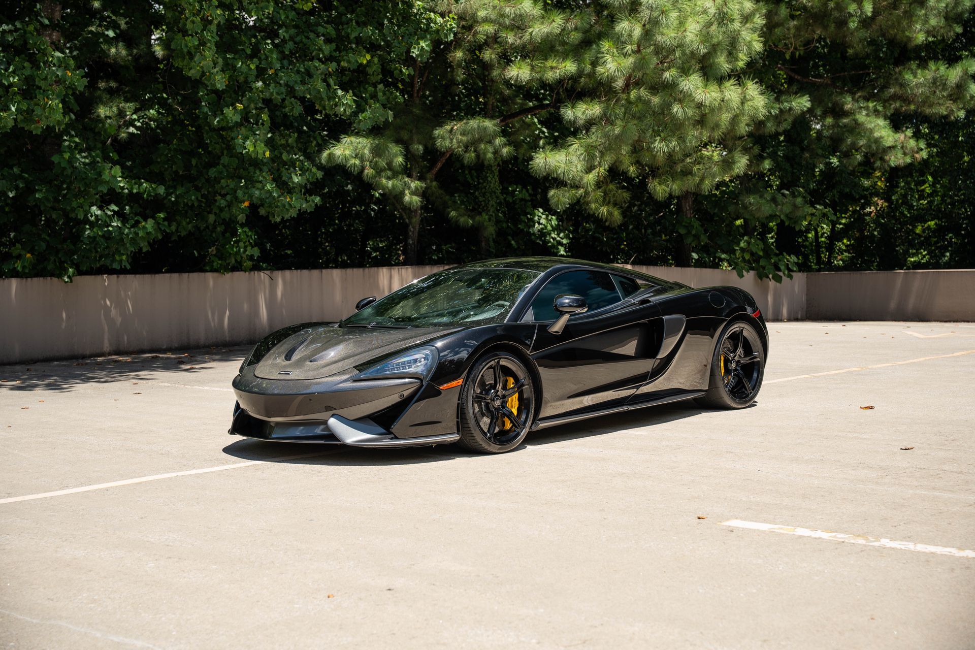 Black McLaren sports car parked on a paved lot with a tree background.