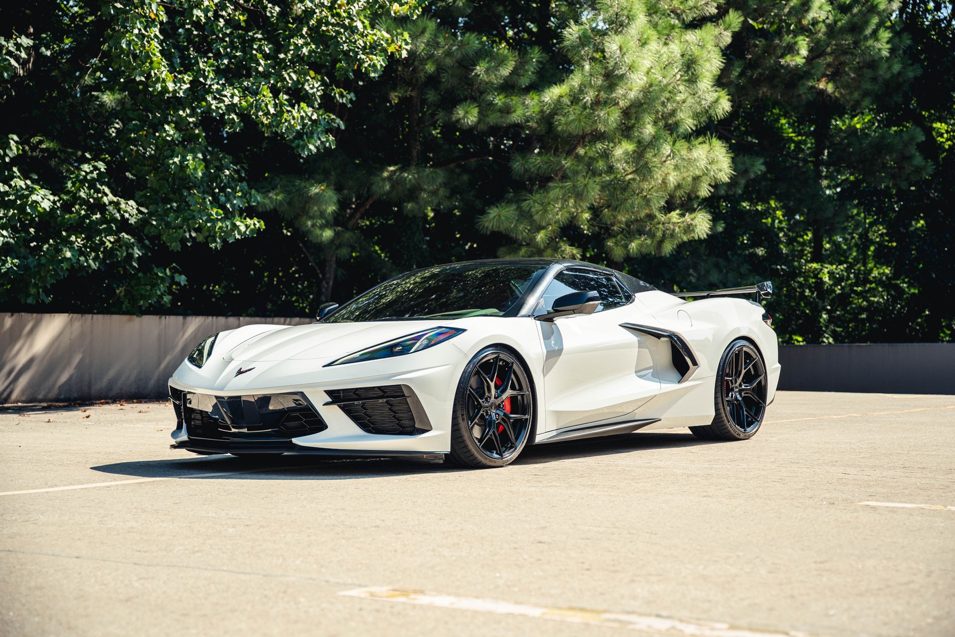 White sports car parked in a parking lot, with a black roof and black wheels, trees in the background.