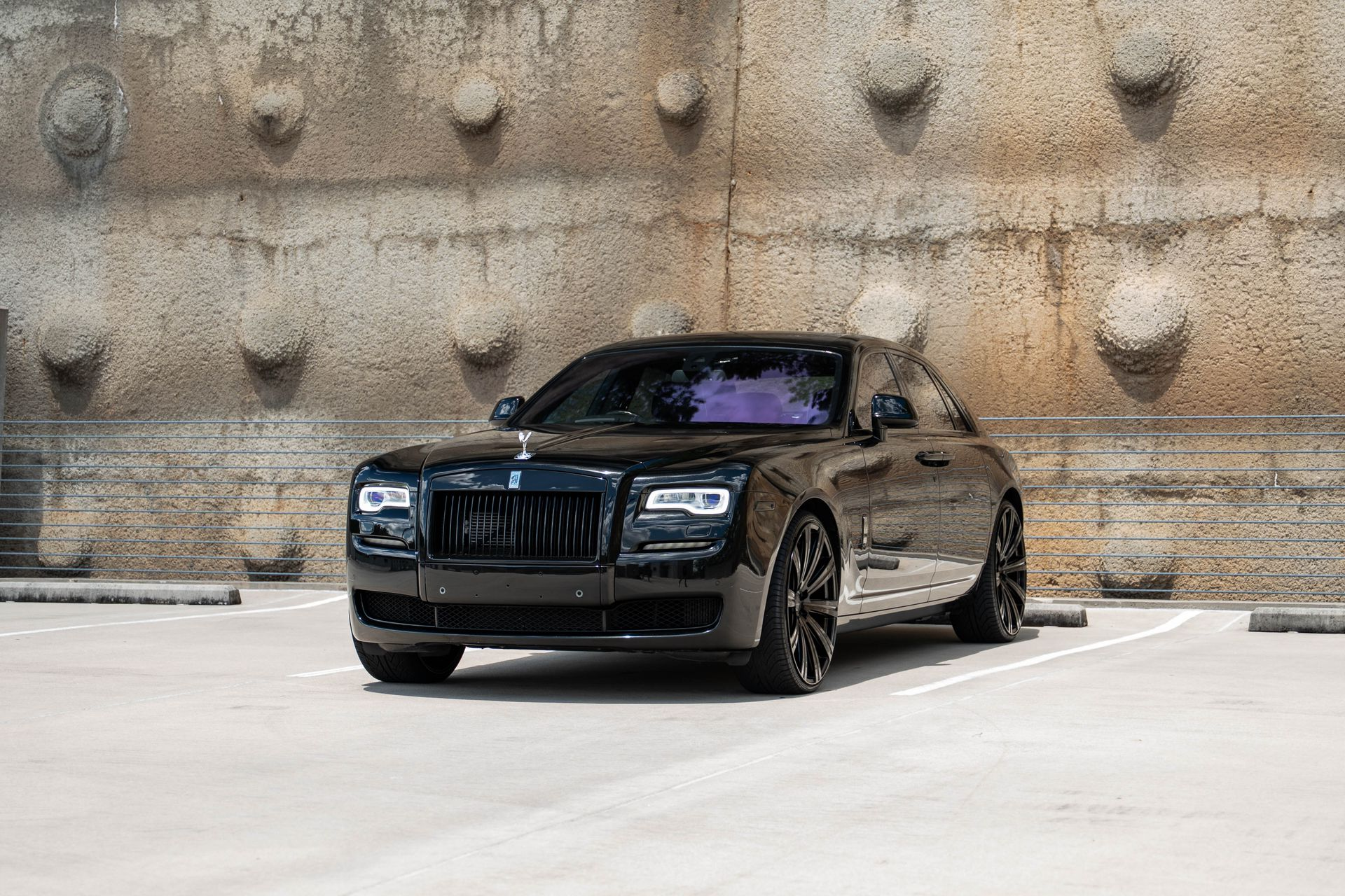 Black Rolls-Royce coupe parked in front of a textured concrete wall; sunny day.