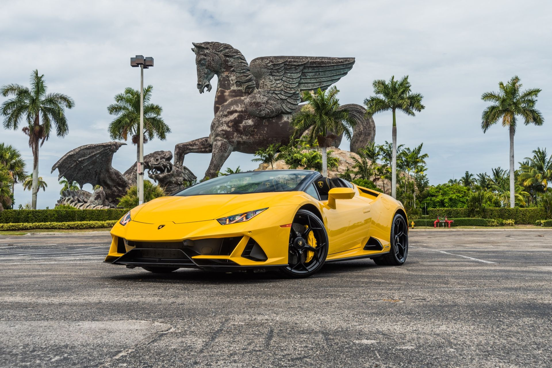 Yellow Lamborghini convertible parked in front of a large horse statue, under a cloudy sky.