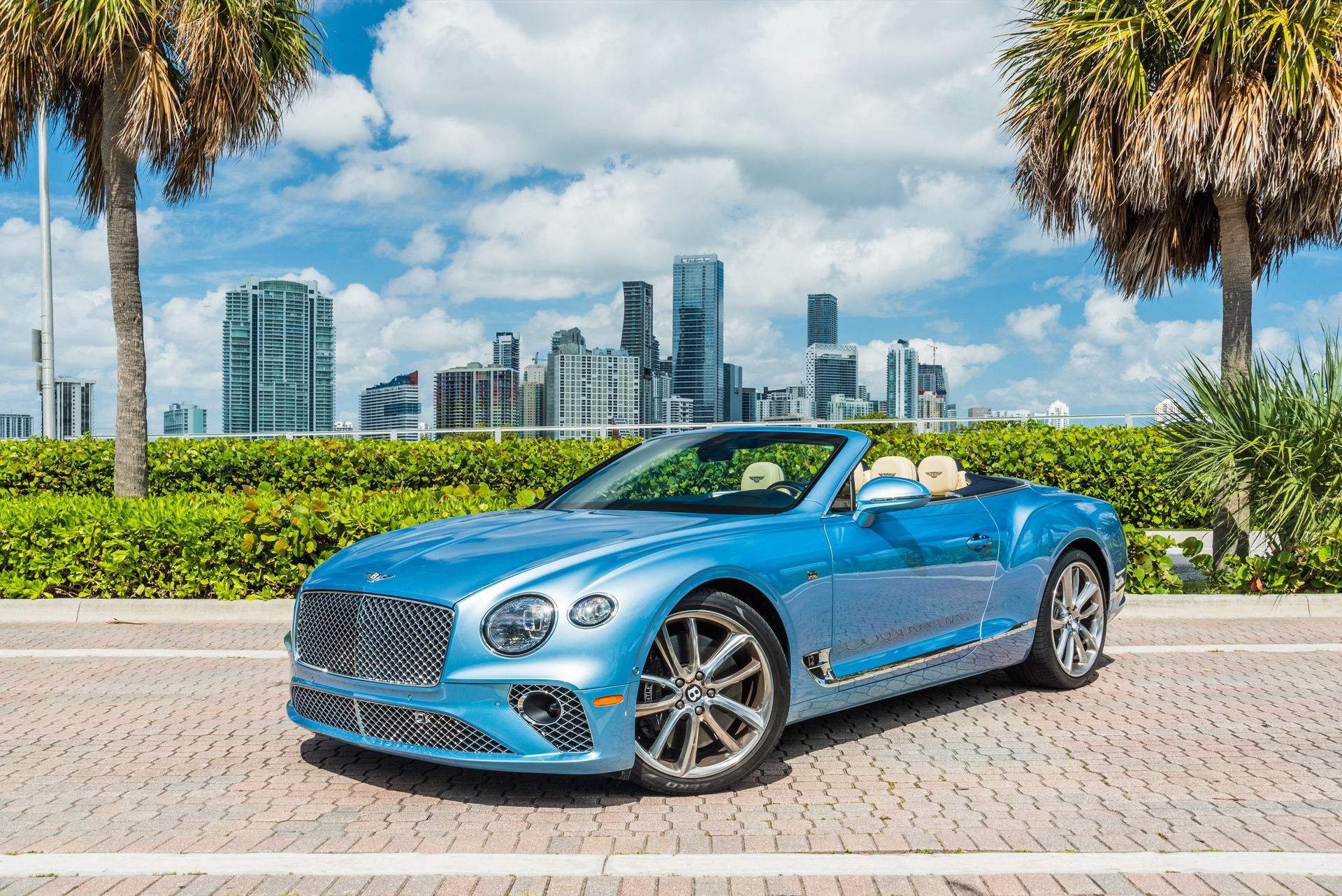Blue Bentley convertible parked on brick, palm trees, city skyline background.