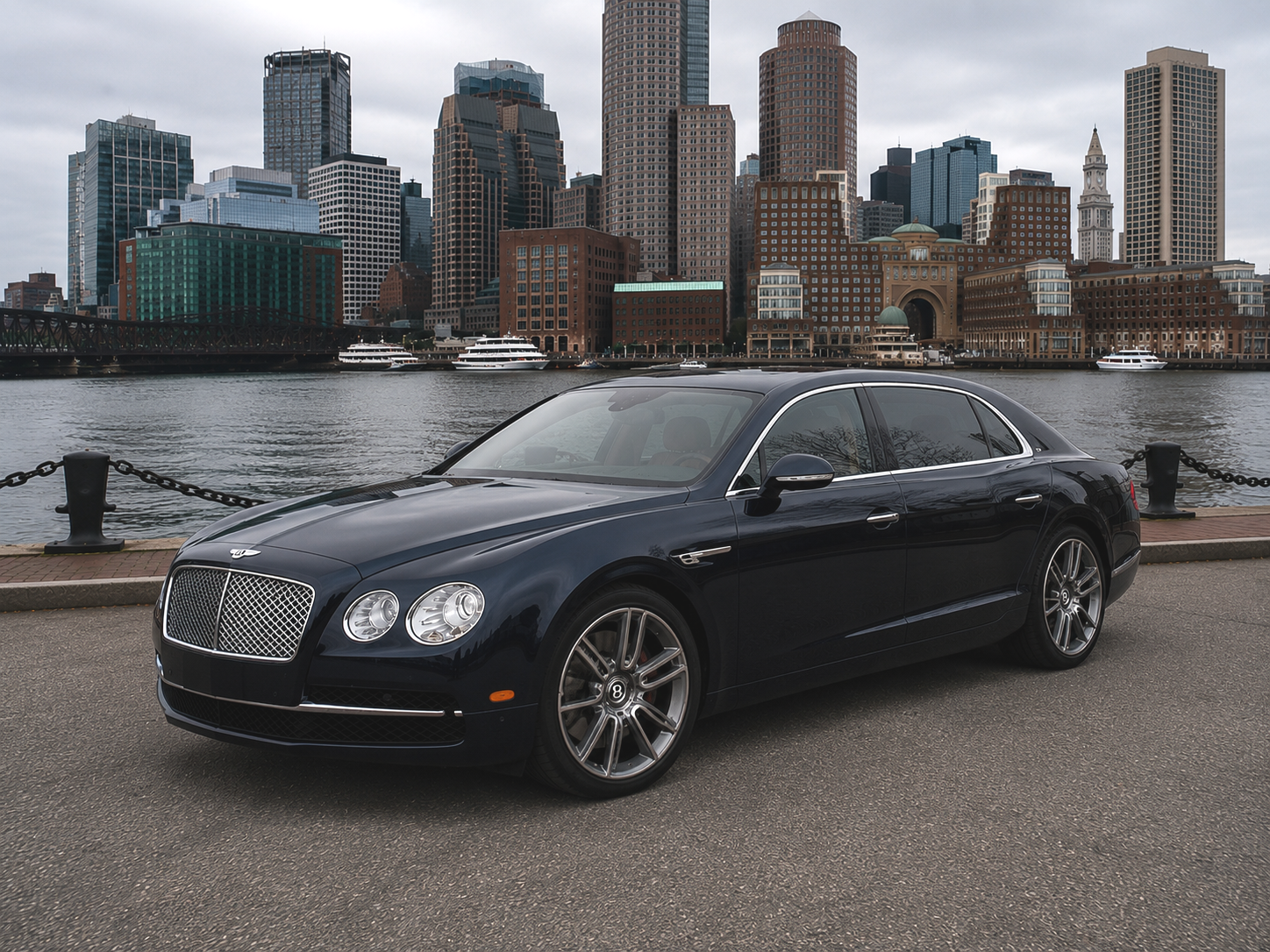 Dark blue Bentley luxury sedan parked by a waterfront with a city skyline in the background