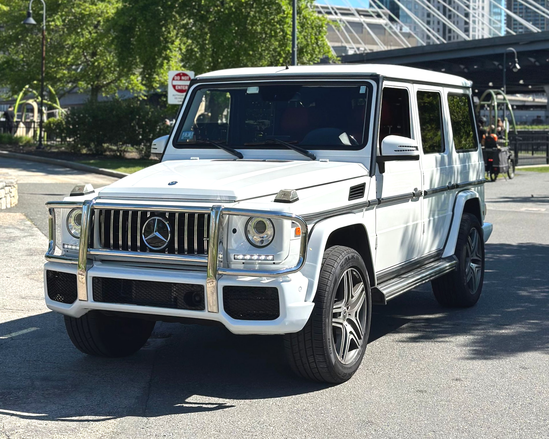 White Mercedes-Benz G-Class SUV parked on a sunny street near a building and trees
