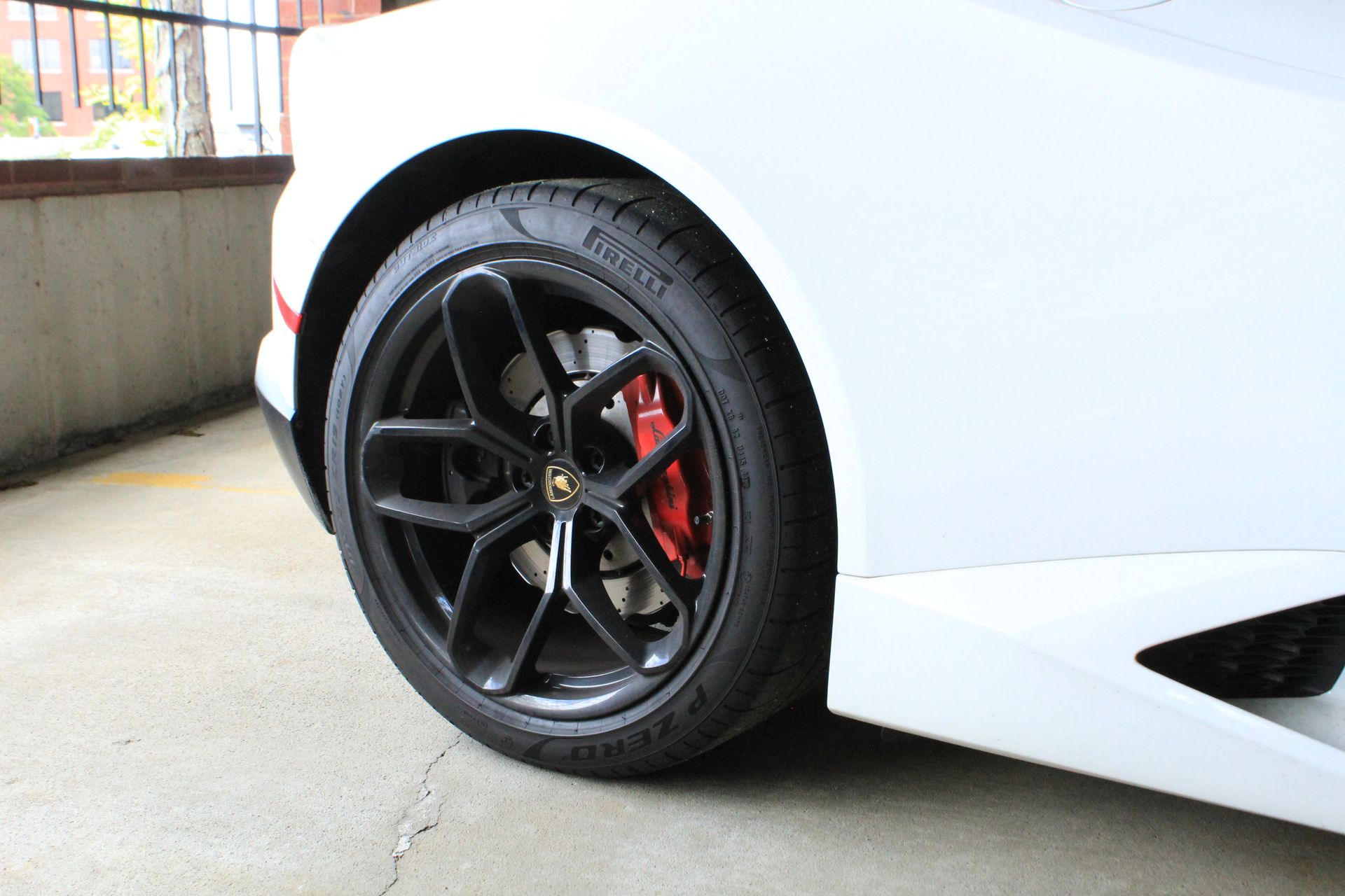 Close-up of a white sports car’s black wheel with red brake caliper, parked by a concrete wall.