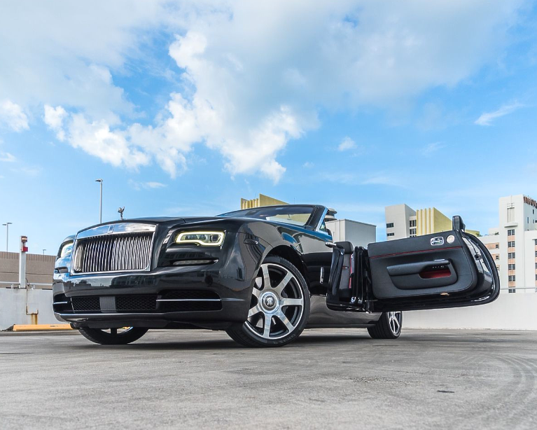 Black Rolls Royce convertible with doors open, parked on a rooftop against a cloudy sky.
