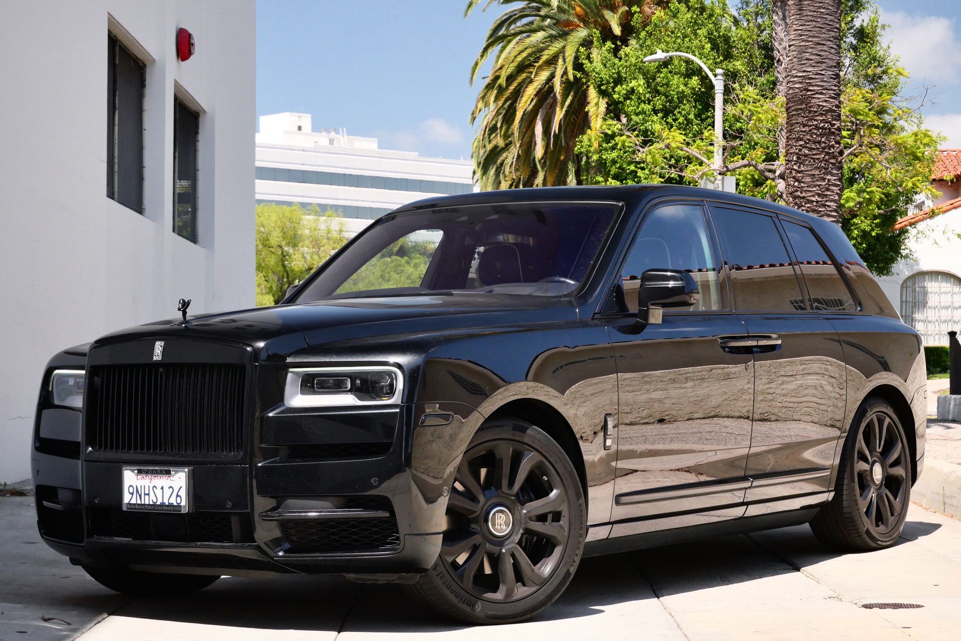 Black Rolls-Royce Cullinan SUV parked on a city street. Black wheels, grille, and trim. Bright sunlight.