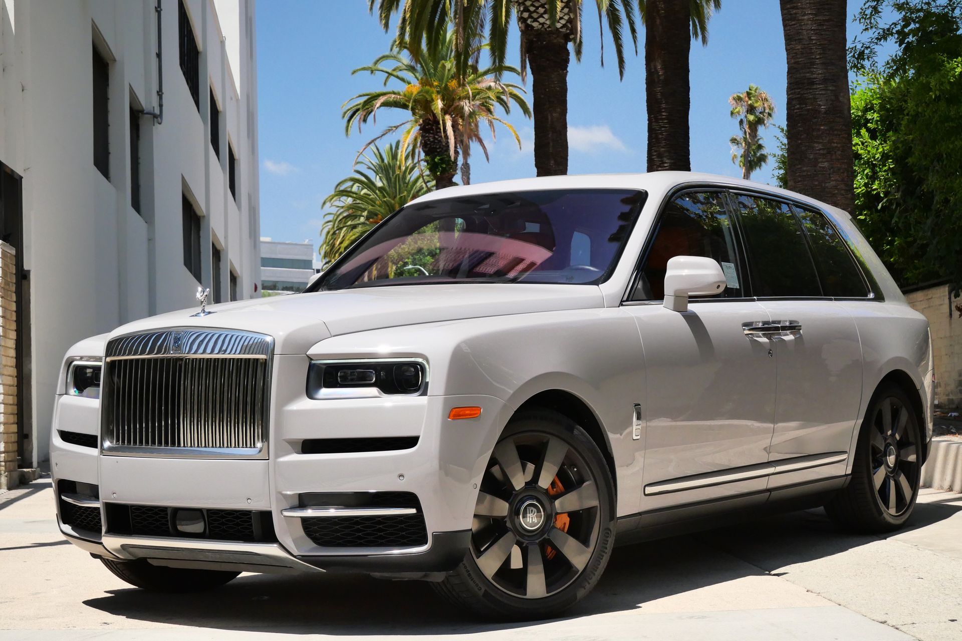 Silver Rolls-Royce Cullinan SUV parked in front of a white building on a sunny day with palm trees.