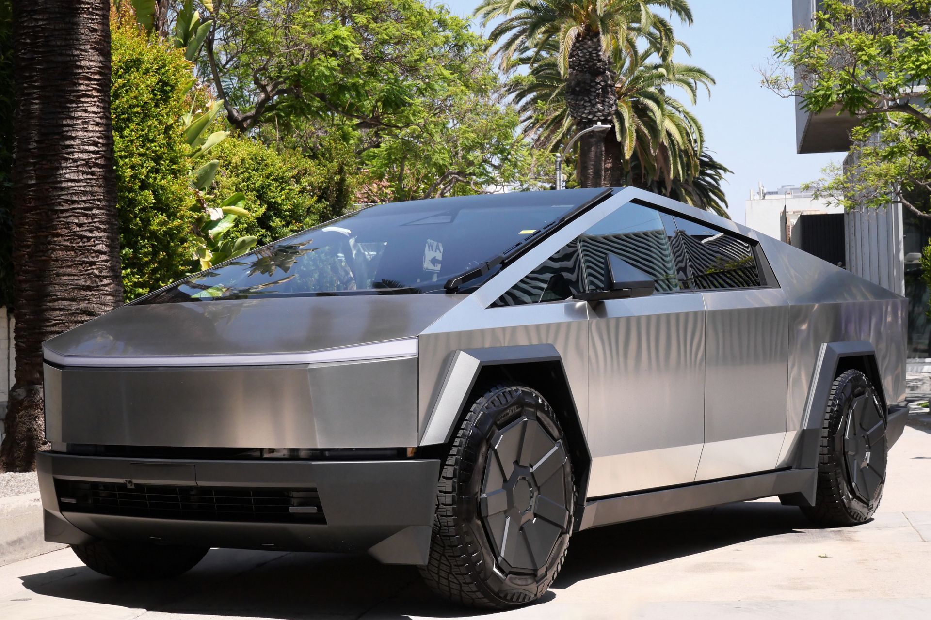 Tesla Cybertruck, silver, parked on street, black tires, in front of palm trees and a building.