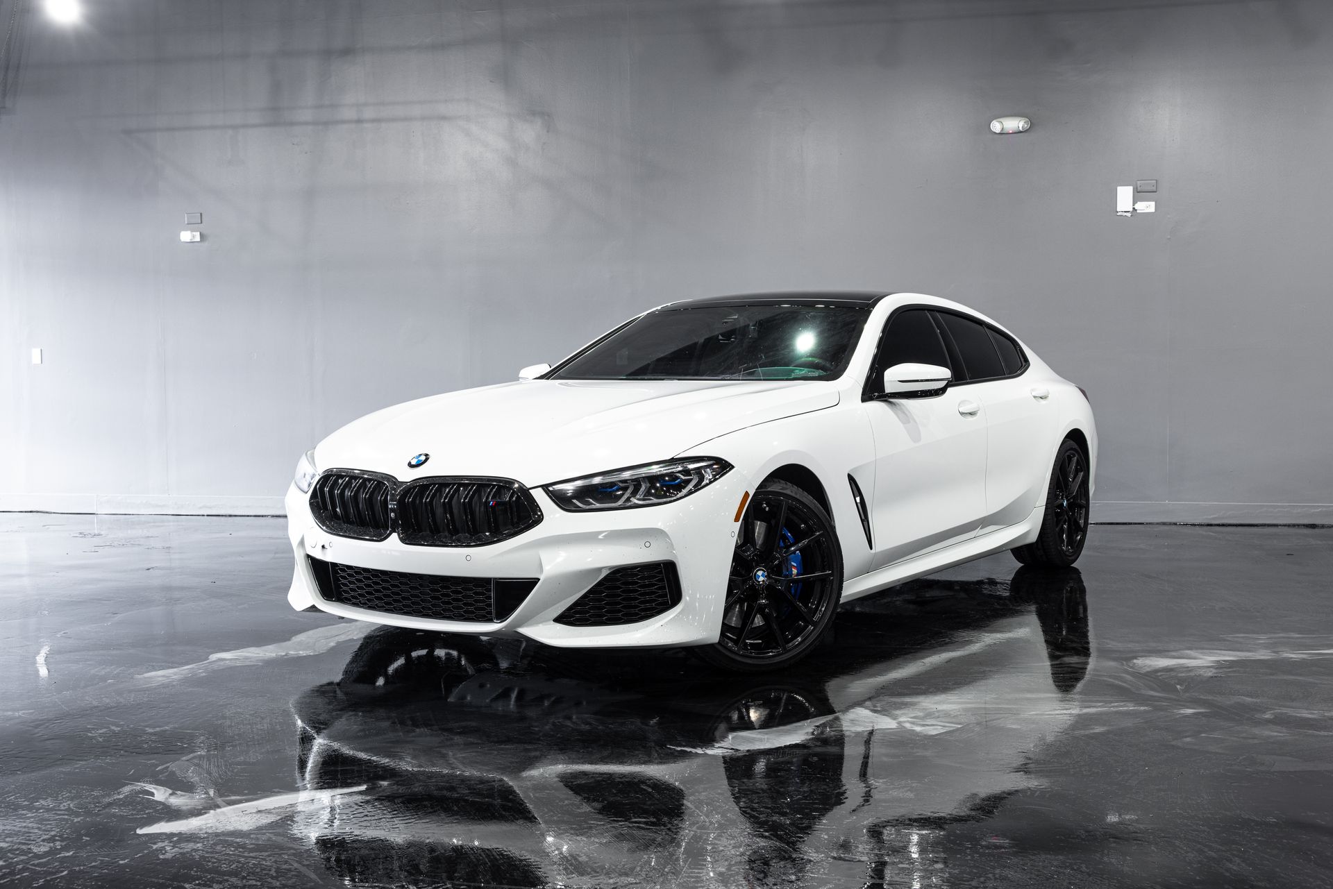 White BMW 8 Series coupe with black accents parked in a studio with a reflective floor.