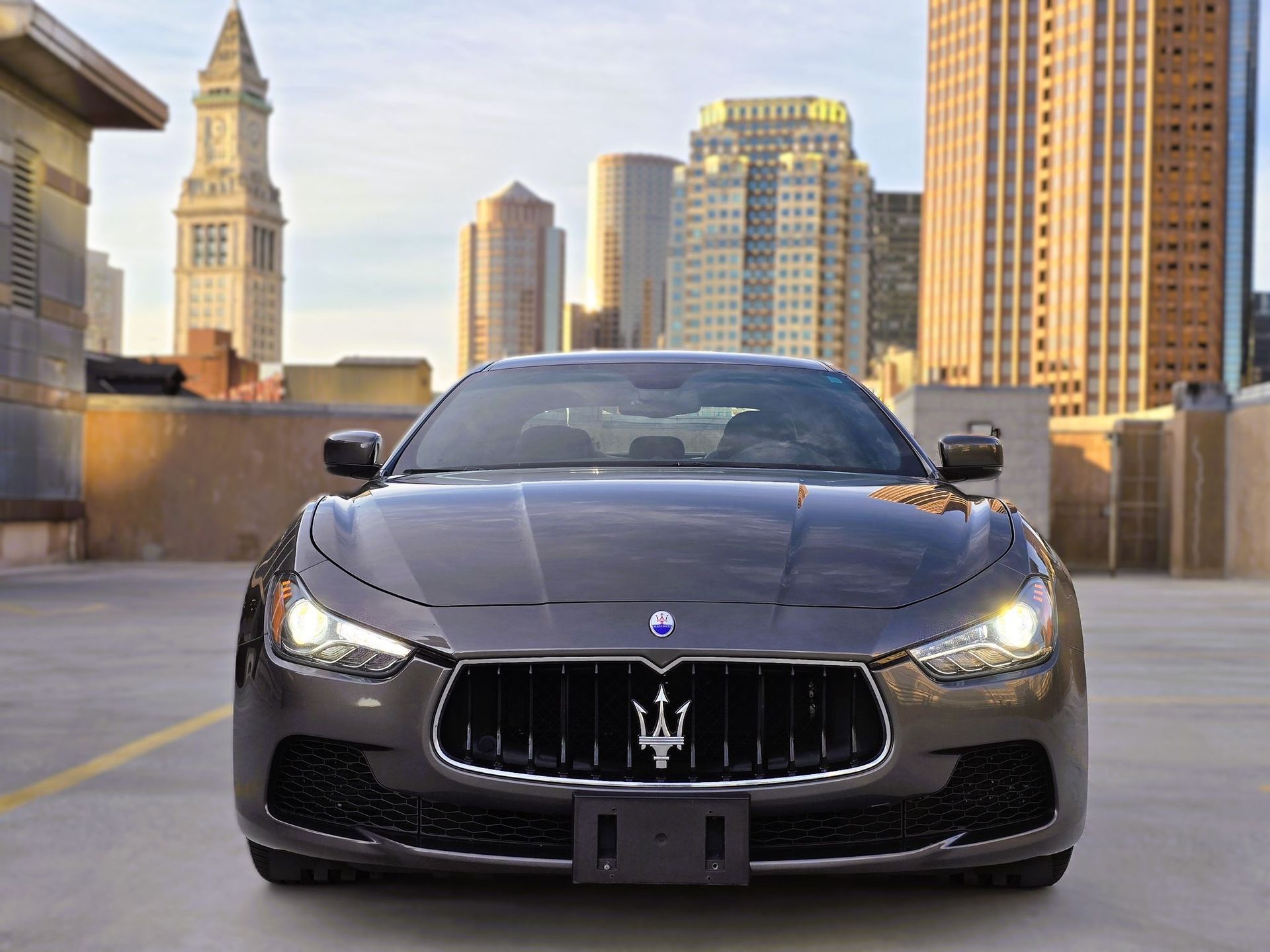 Front view of a gray Maserati sports car parked on a rooftop with a city skyline at sunset