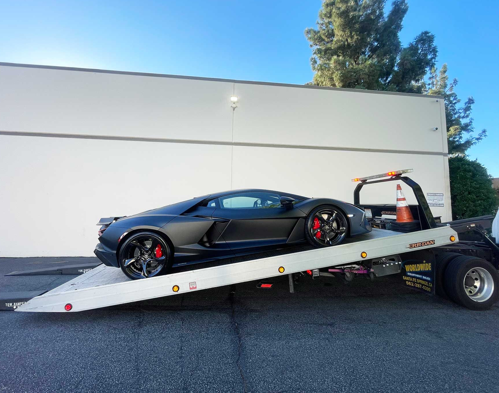 Black Lamborghini loaded on a flatbed tow truck in front of a white building.