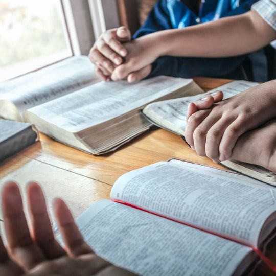 Several people sit around a wooden table, hands resting on open books while others are clasped in prayer.