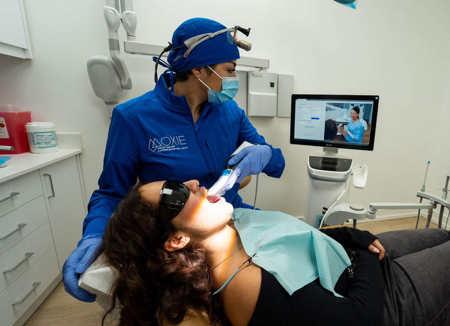 Dentist and assistant examining patient's teeth in a dental office. Both wear masks, patient reclined.