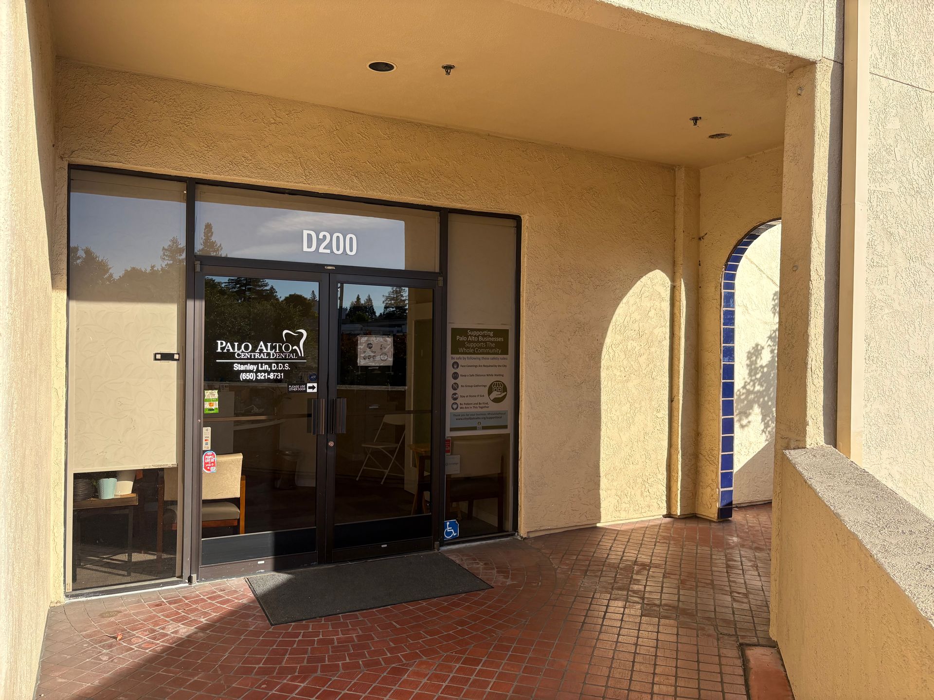 Building entrance with glass doors, tan walls, and a brick walkway under an overhang.
