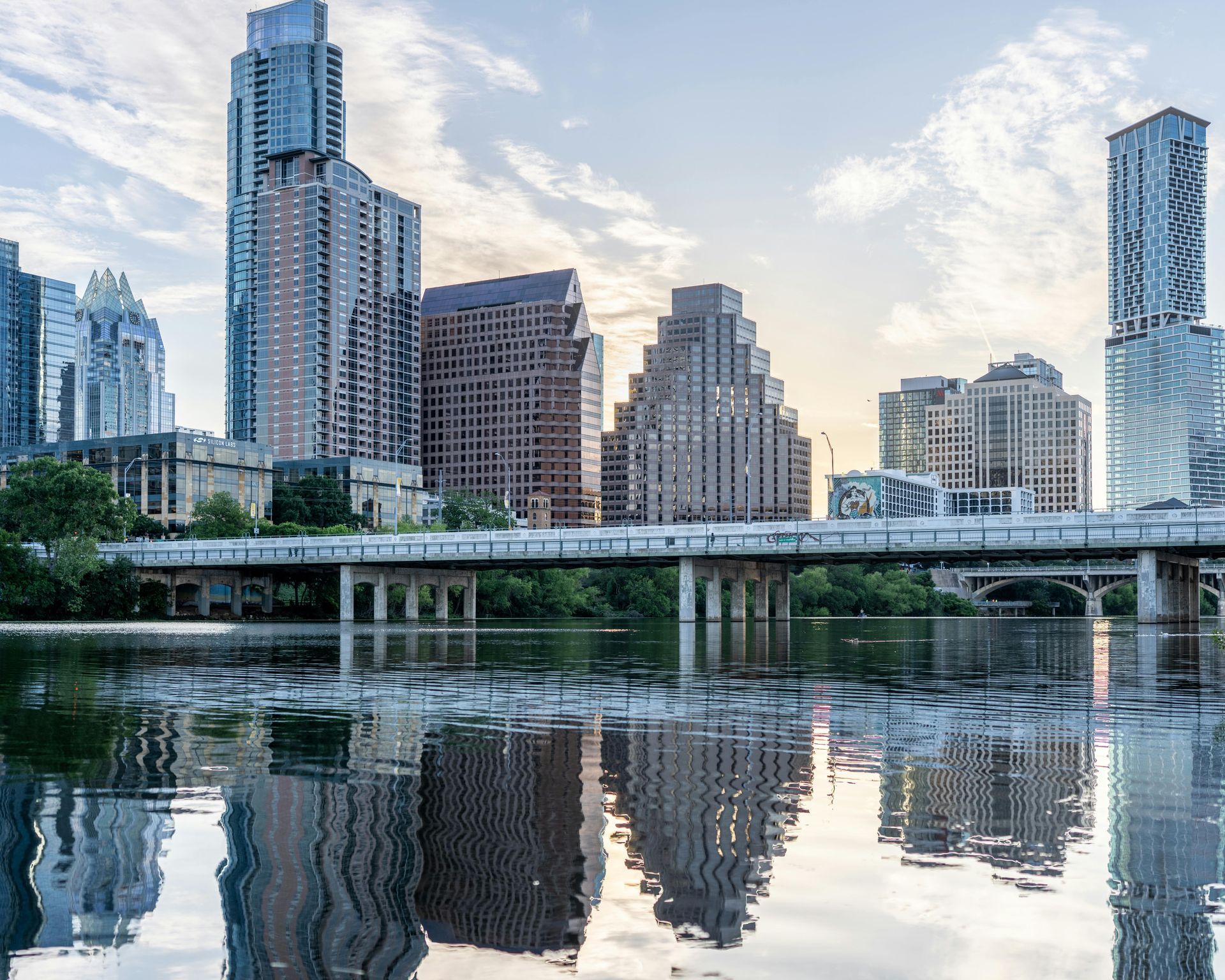 Modern high-rise buildings reflected in a calm river beneath a bridge at dusk
