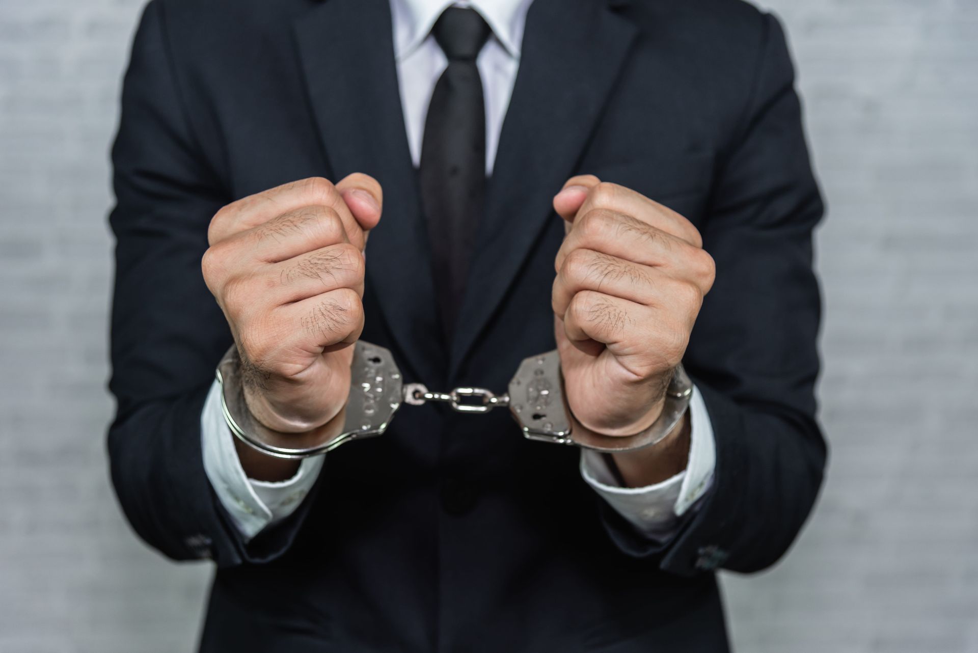 Person in a black suit with handcuffs on their wrists against a gray background
