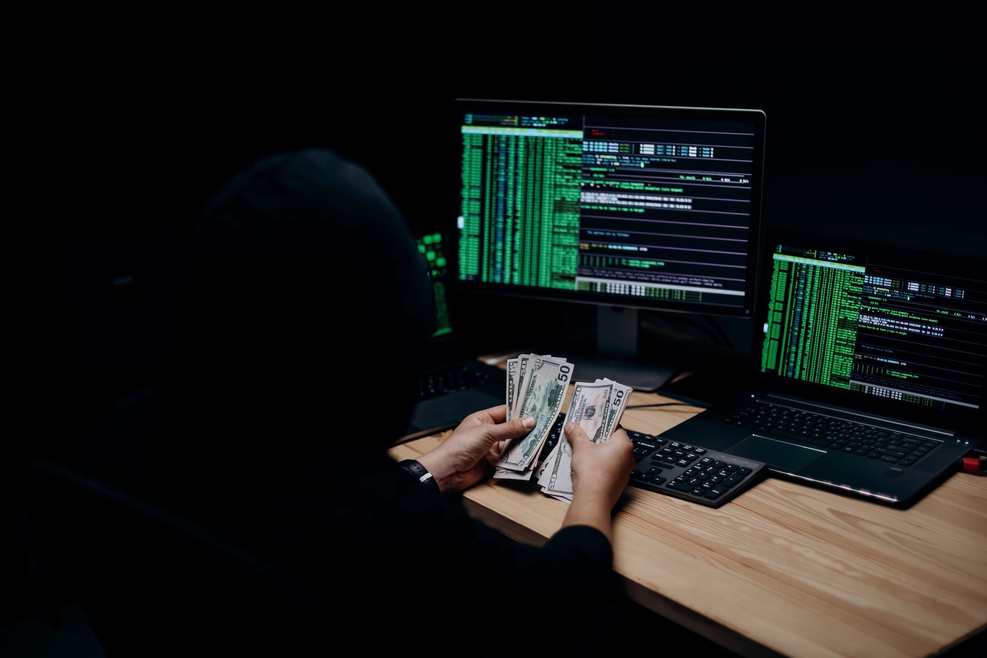 Hooded person counting cash at a desk with two monitors showing green code in a dark room