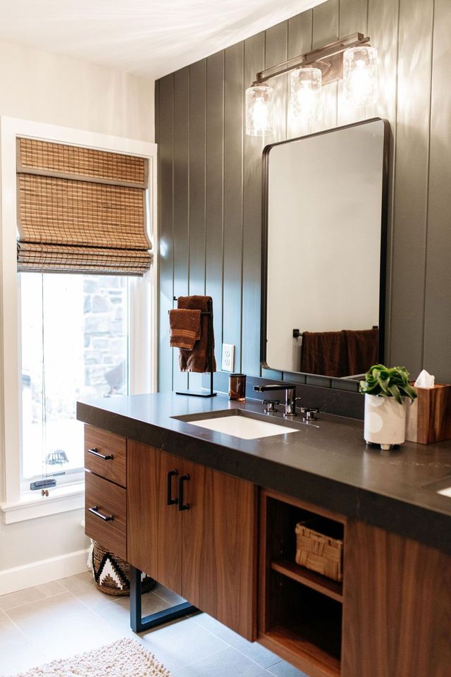 Modern bathroom vanity with dark wood cabinets, black countertop, mirror, window with woven shade, and potted plant