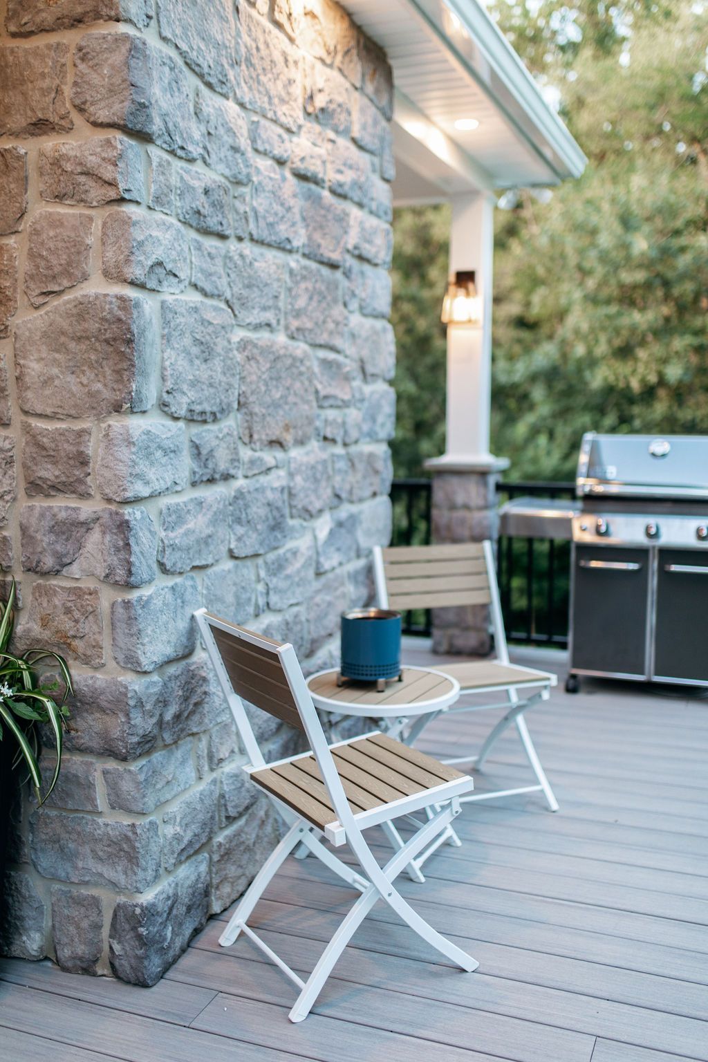 Stone patio with two white folding chairs and a small table beside a brick house and grill.