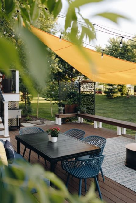 Outdoor patio with black table and chairs under a yellow shade sail, surrounded by greenery