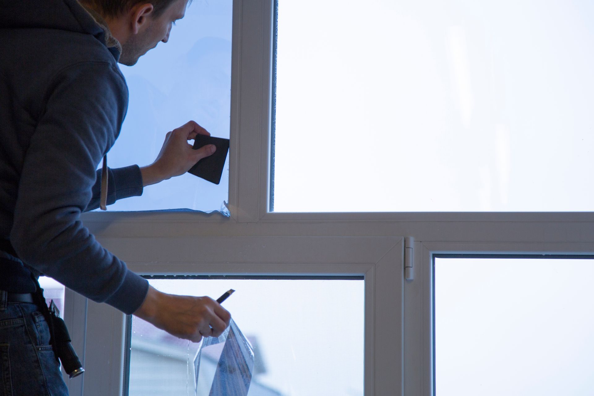 Person applying window tint, using a squeegee and knife, on a white-framed window.