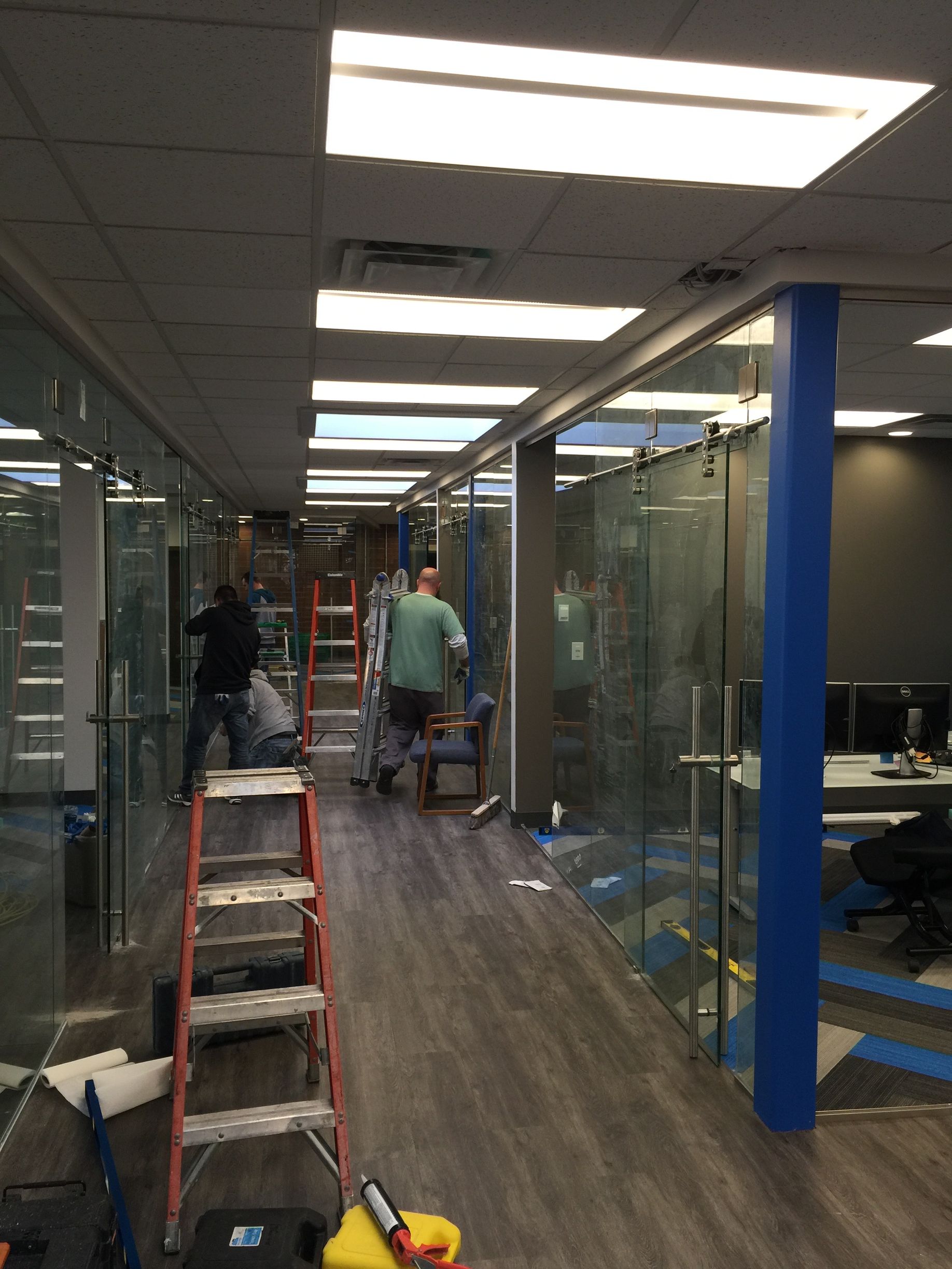 Workers installing glass office walls in a commercial space with dark flooring and blue accents.