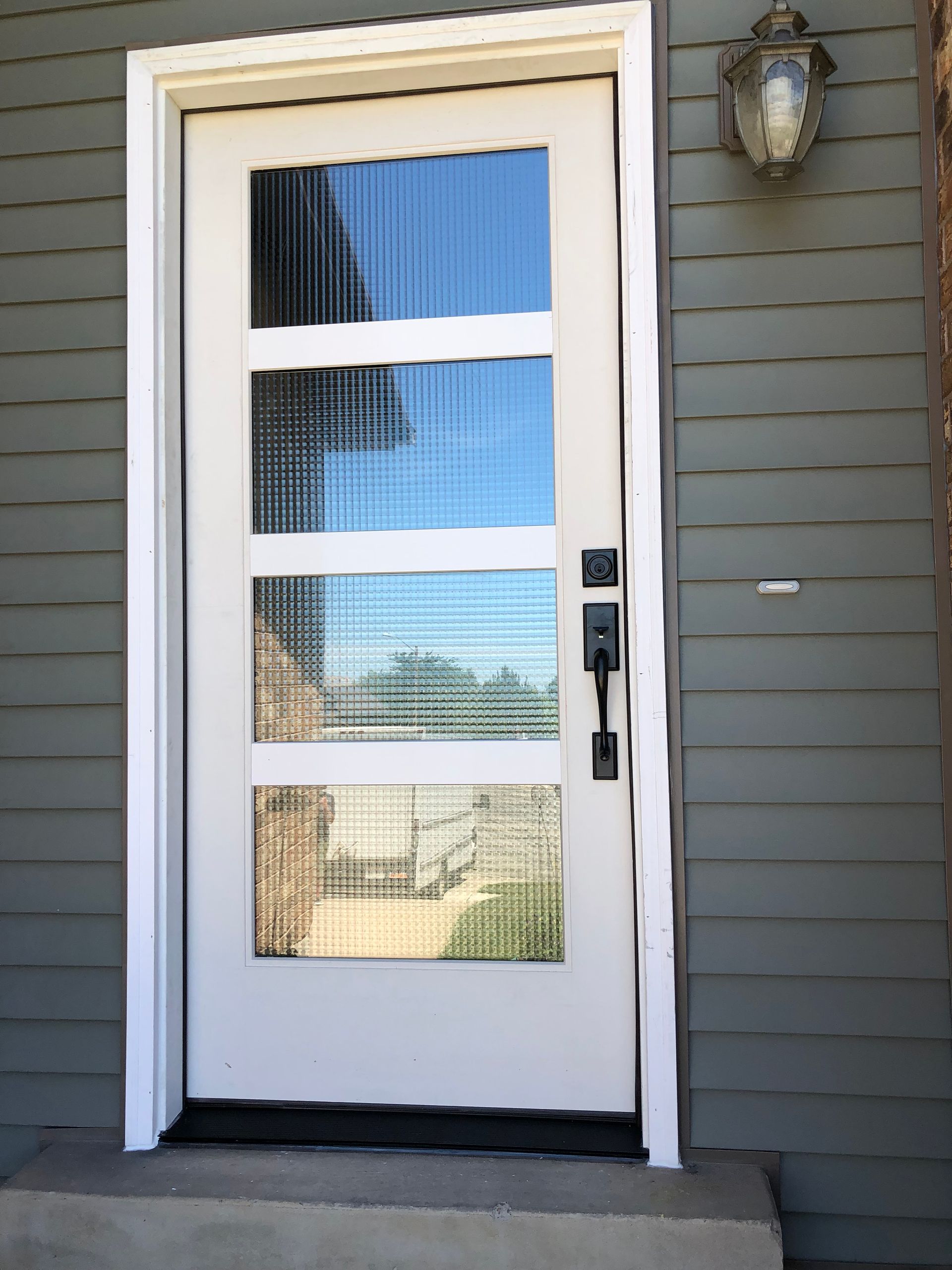 White front door with four glass panels, black hardware, set in a gray-sided building.