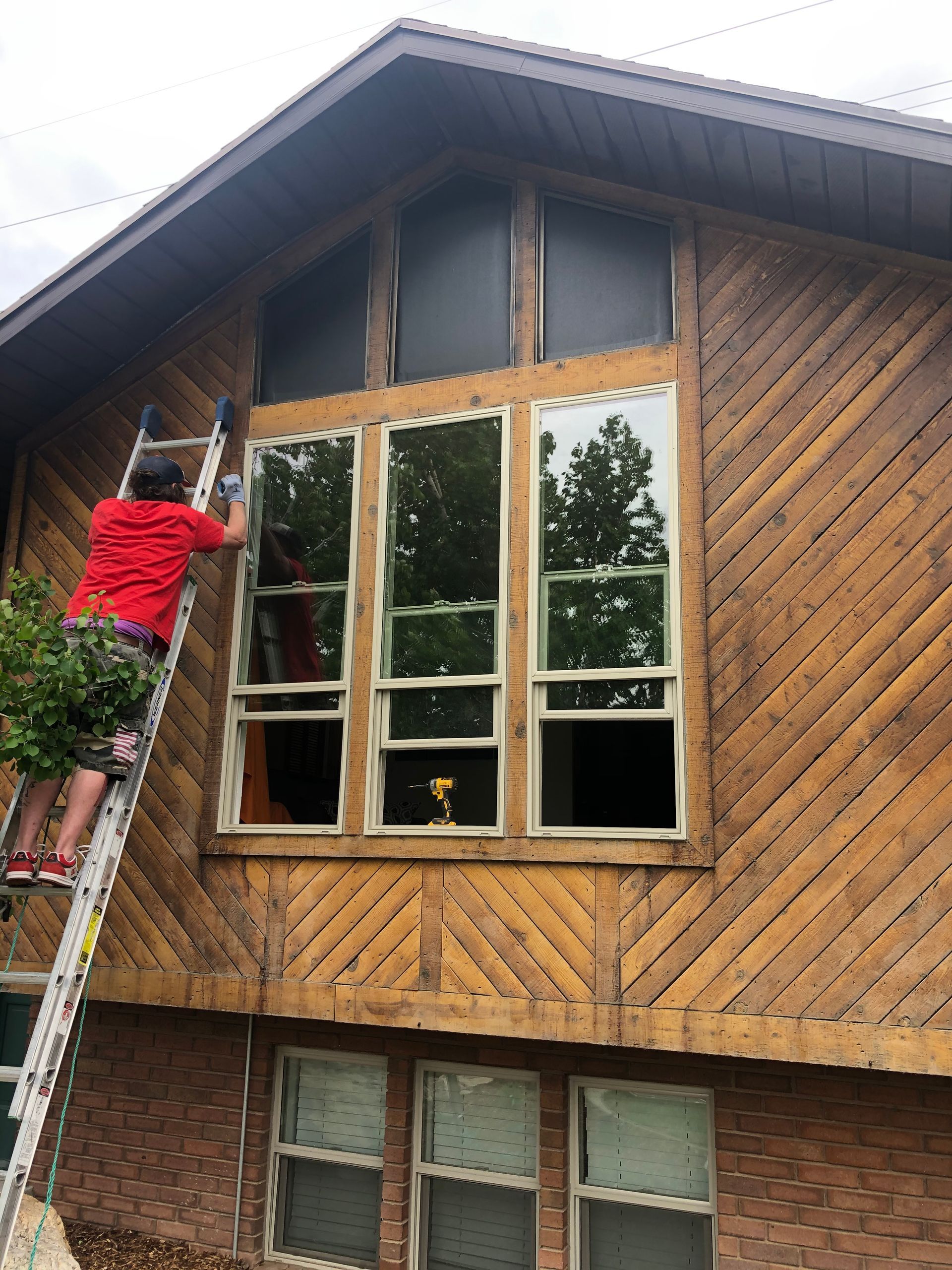 Person on ladder cleaning large windows on a wood-paneled house exterior.
