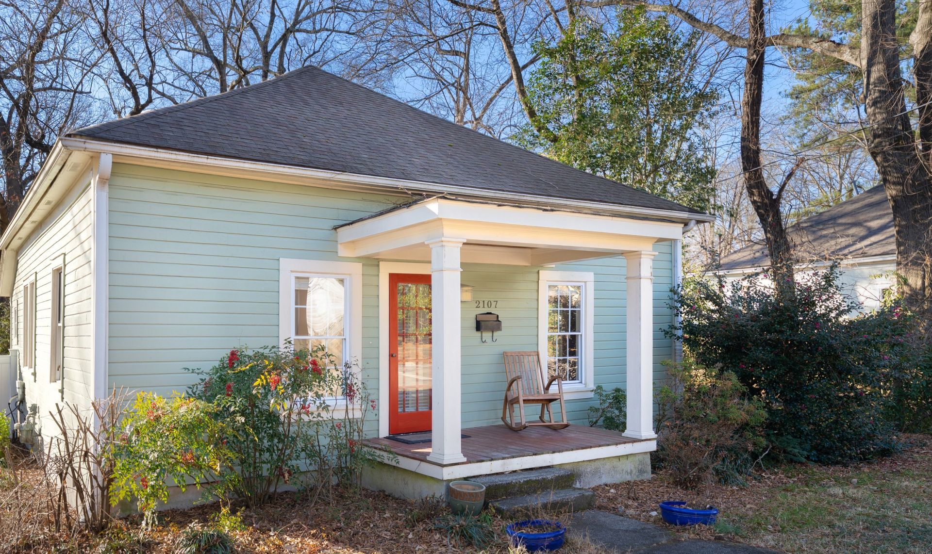 A light green, single-story house with a white-pillared porch, a bright orange front door, and trees in the background.