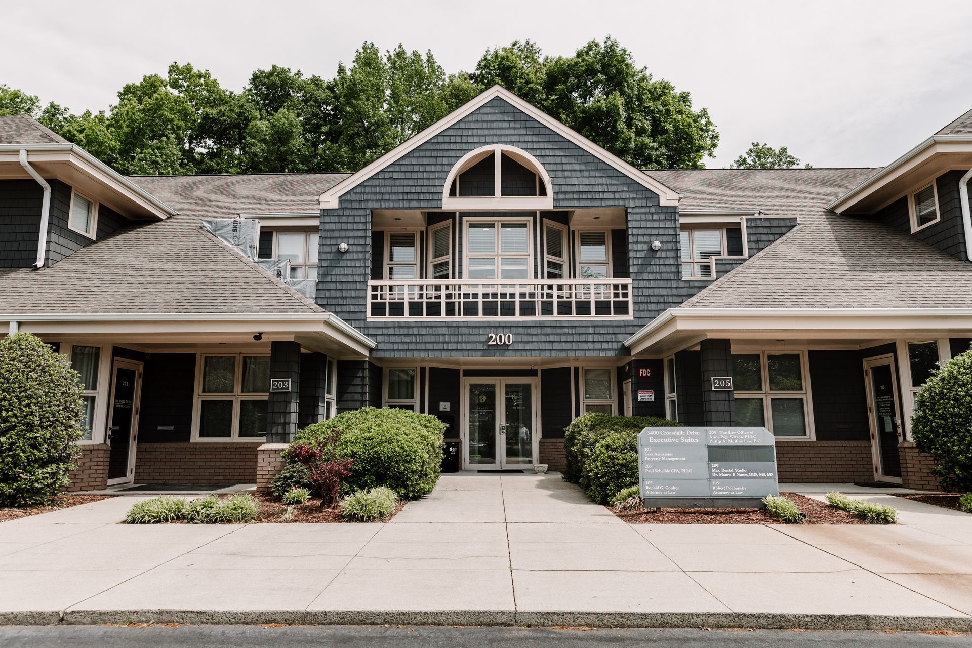 A two-story commercial building with dark gray shingles, beige trim, a central balcony, and a directory sign in front.