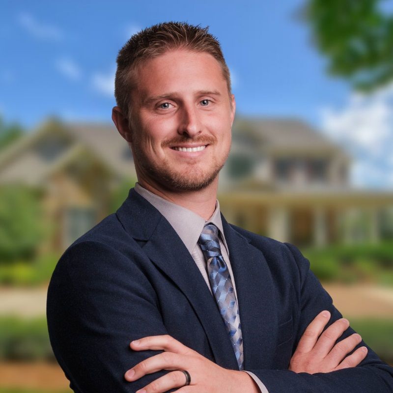 A smiling professional in a navy suit and patterned tie poses with arms crossed in front of a blurred residential home.
