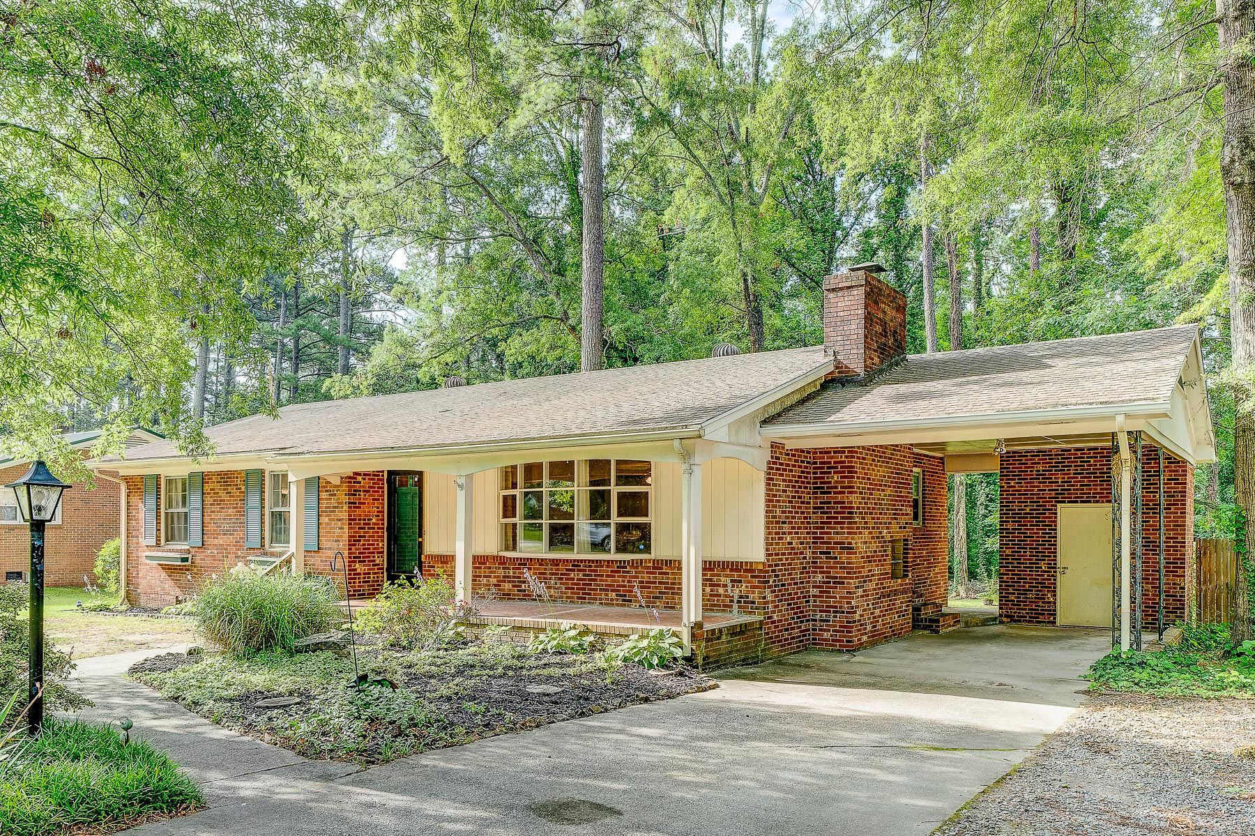 A tan, single-story house with a brick accent, covered porch, and metal roof, nestled among tall green trees.