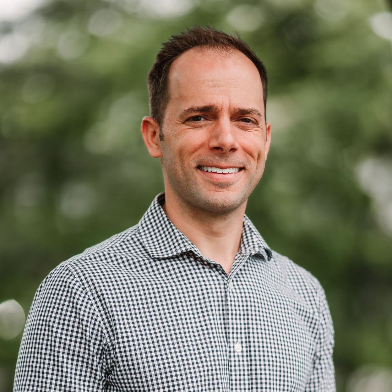 A smiling man in a blue and white checkered shirt standing outdoors against a blurred green background.