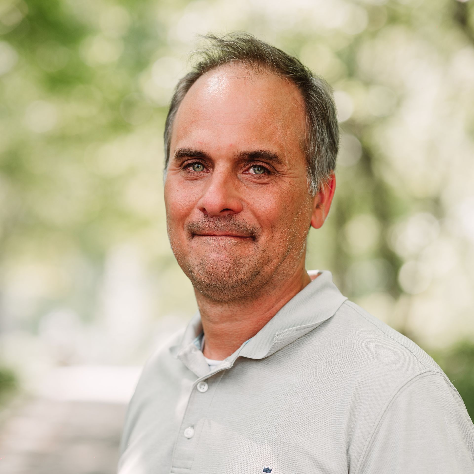 A person with short, thinning hair wearing a light gray collared shirt, smiling gently in an outdoor, blurred background.
