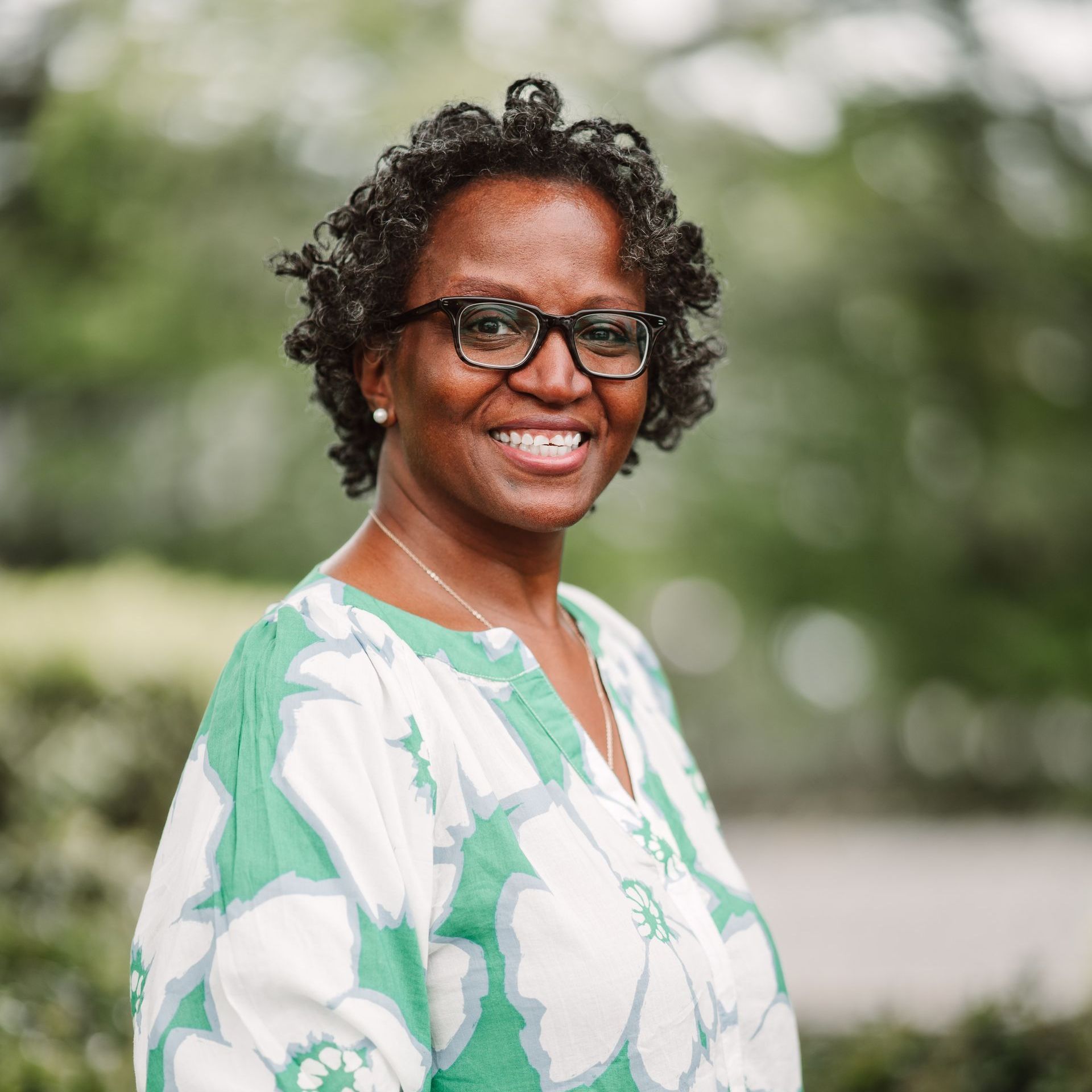 A person with short, curly hair and glasses smiles while wearing a green and white floral-print top in an outdoor setting.