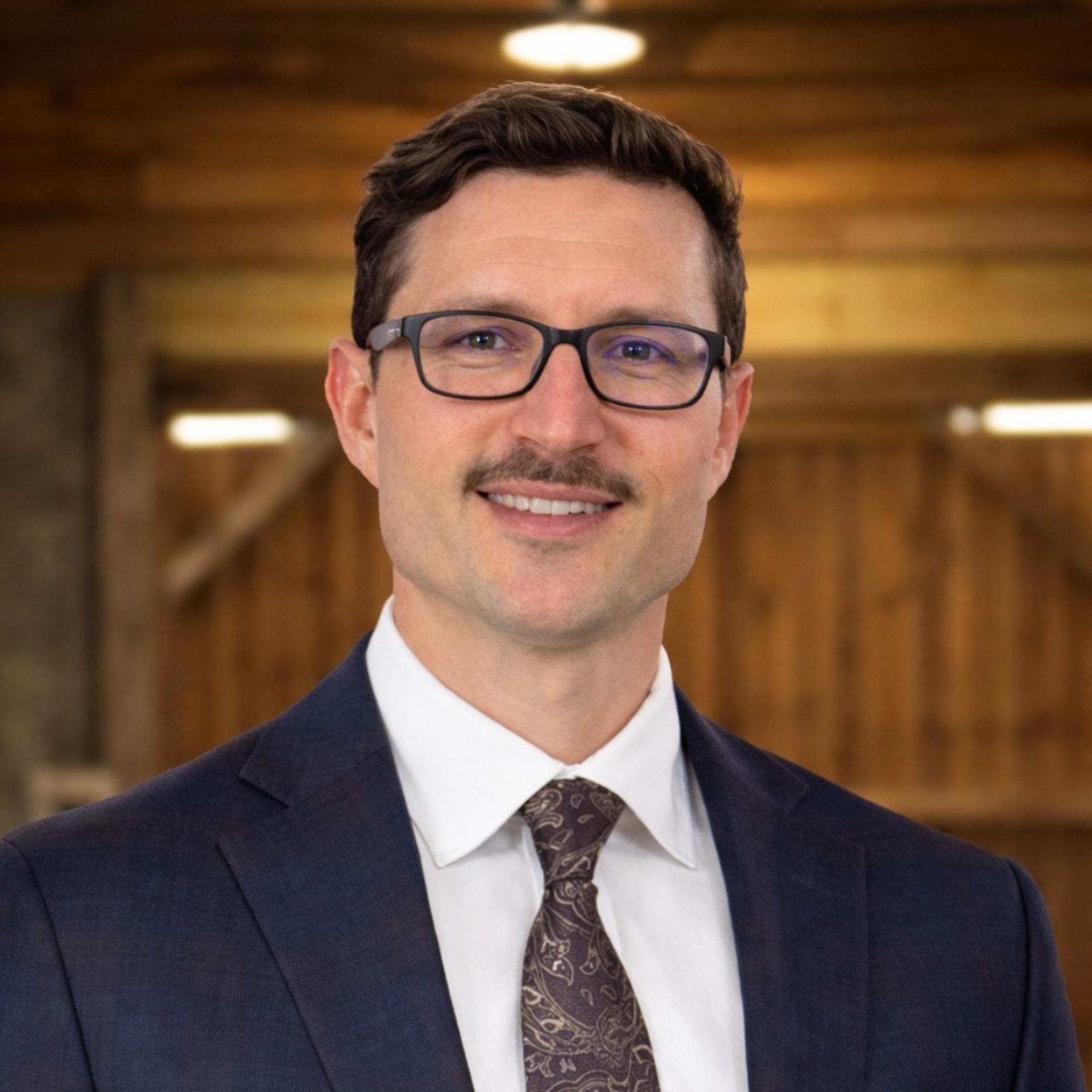 Headshot of a smiling person wearing glasses, a dark blue blazer, white shirt, and patterned brown tie against a wood wall.