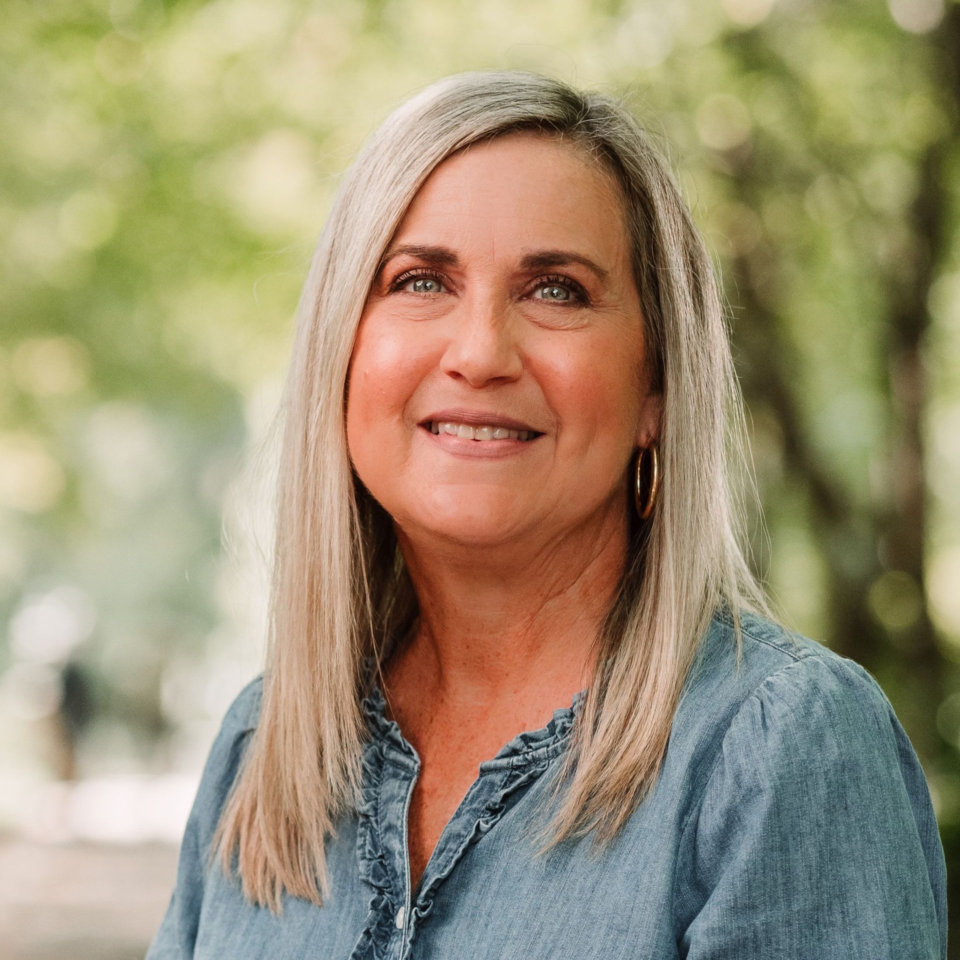 A person with long blonde hair and a friendly smile, wearing a blue denim shirt against a soft, blurred outdoor background.