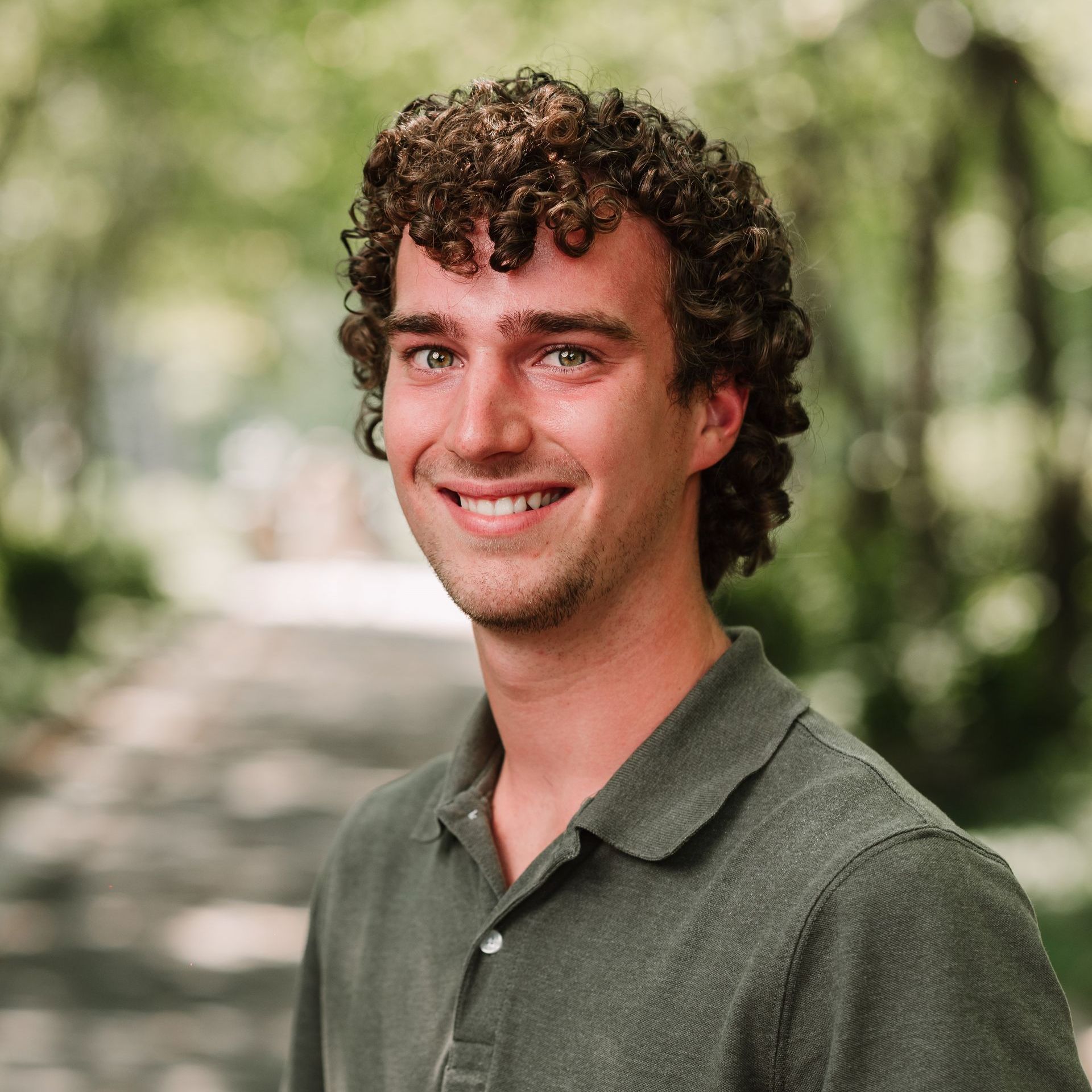 A smiling person with curly hair wearing a green polo shirt, posed outdoors with a soft-focus, leafy background.
