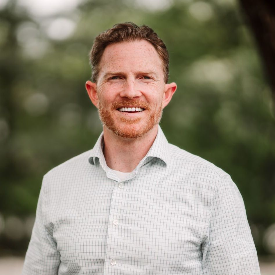 A person with short auburn hair and a beard, wearing a light patterned button-down shirt, smiles outdoors.