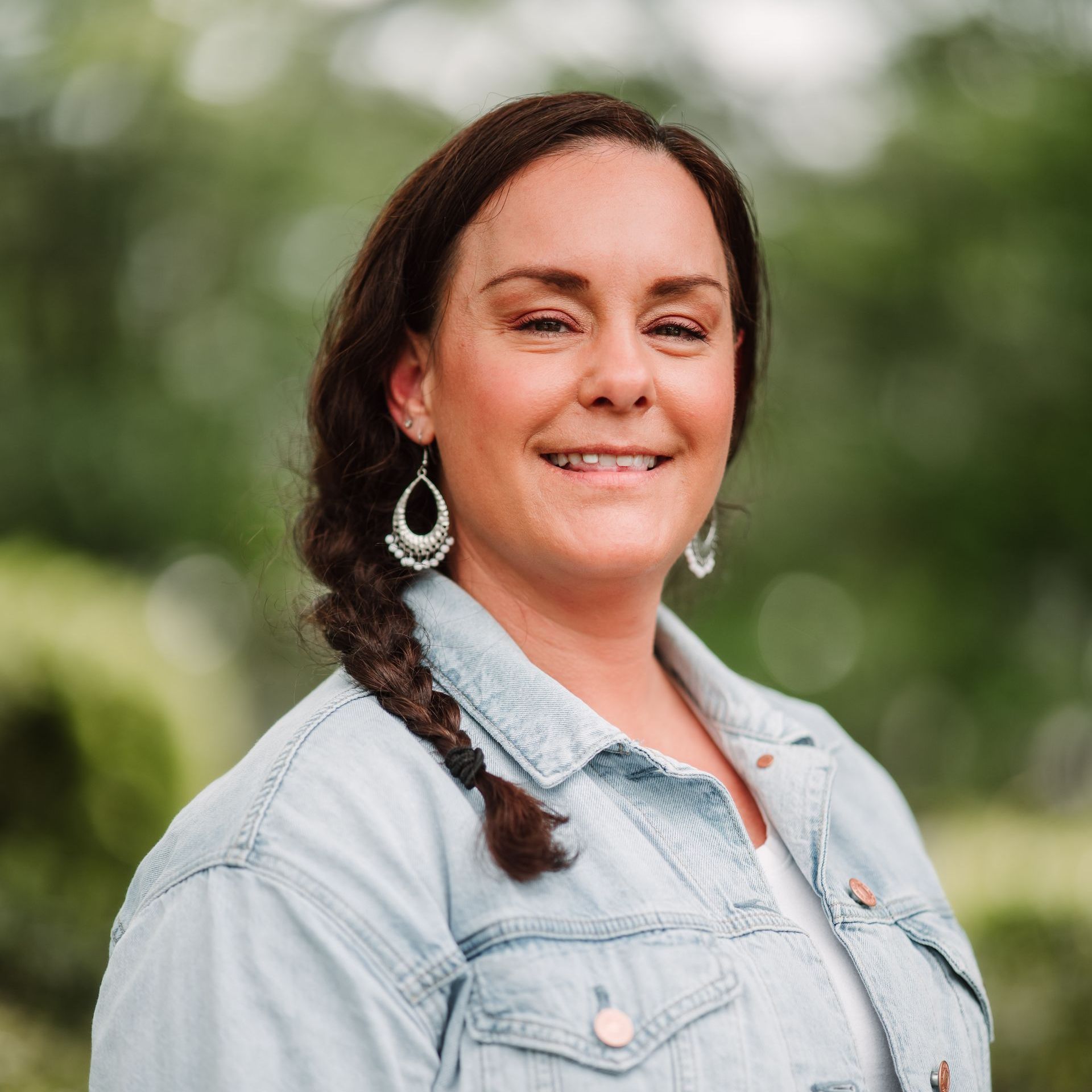 A person with a braided ponytail wearing a light blue denim jacket smiles against a soft, blurry outdoor green background.