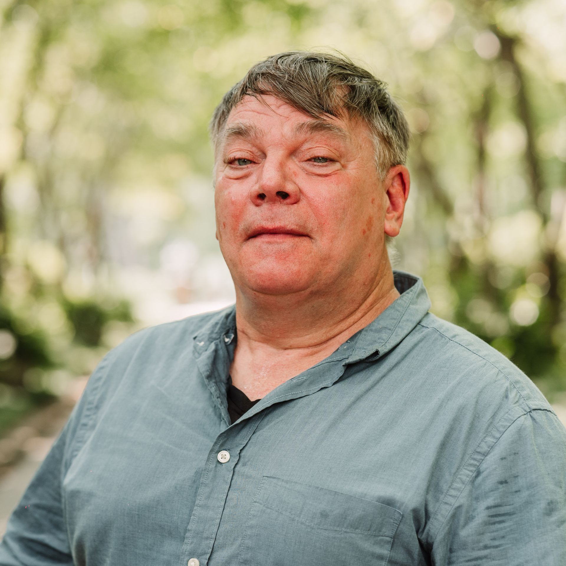 A person with short, tousled hair wearing a blue button-up shirt, posing against a soft-focus, leafy green background.