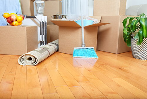 Cardboard boxes, a rolled-up rug, and a blue broom are on a polished wood floor during a home move.