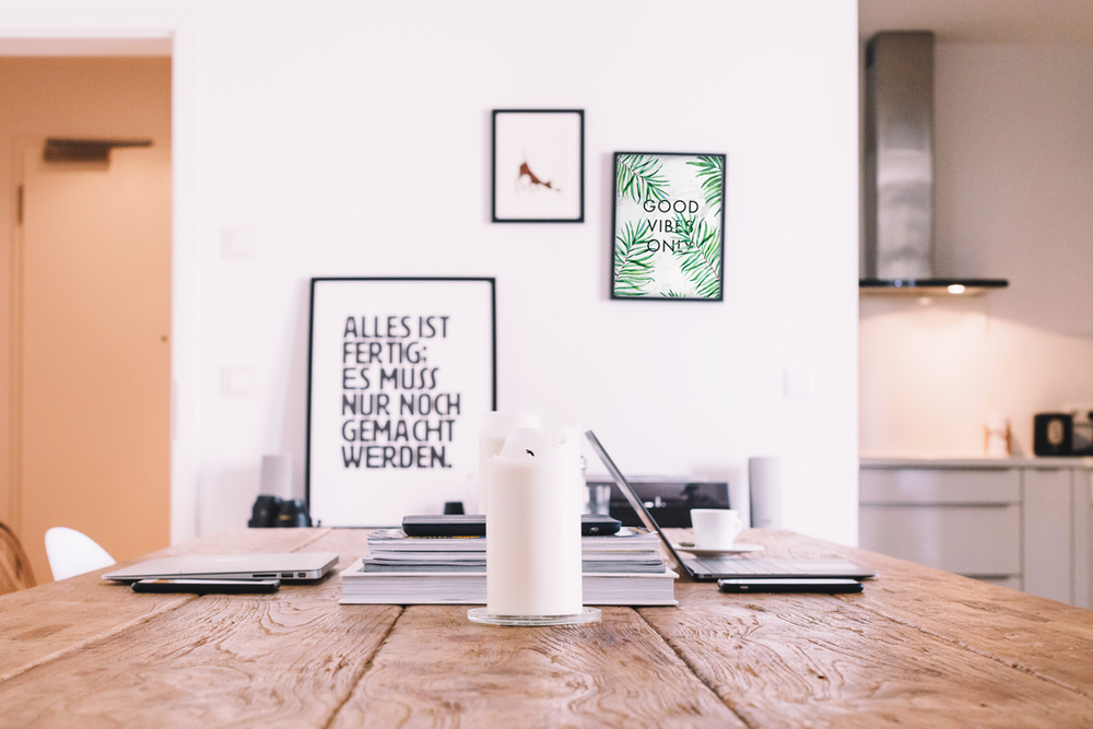 A white pillar candle stands on a wooden desk in a bright, modern room, with framed wall art and a kitchen in the back.